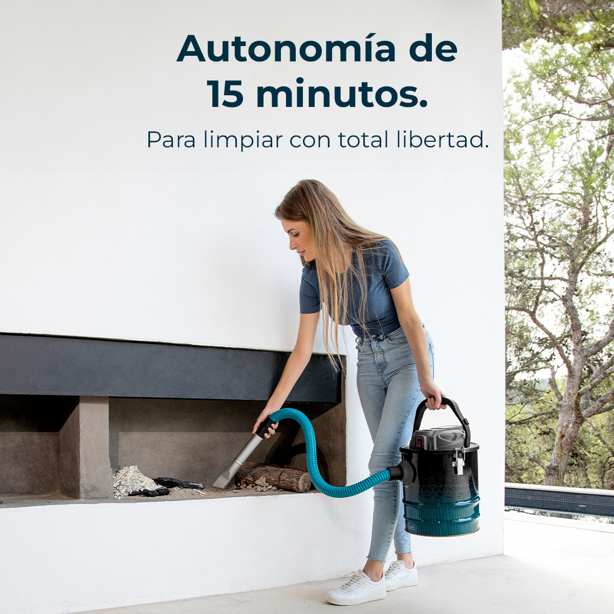 Una mujer aspira una chimenea. Usa una manguera azul conectada a una aspiradora negra y turquesa. El fondo muestra árboles y pared blanca.