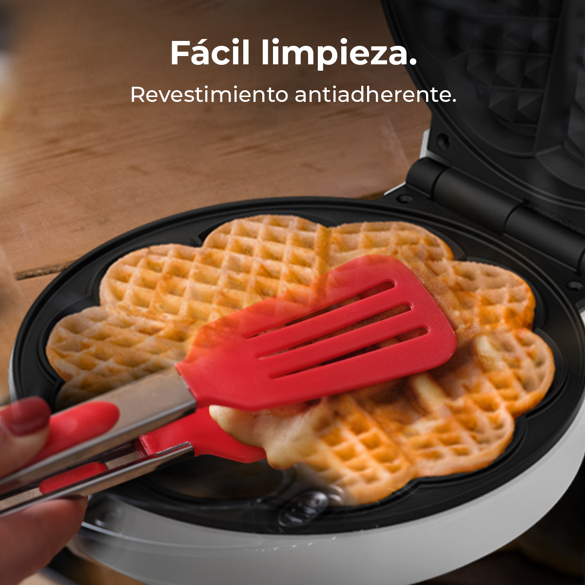 Una macchina per waffle con waffle a forma di cuore. Una spatola rossa rimuove un waffle. Lo sfondo è una superficie di legno.