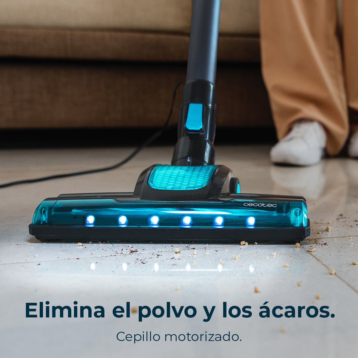 A vacuum cleaner head with blue lights is on a tiled floor with debris. A person stands in the background.