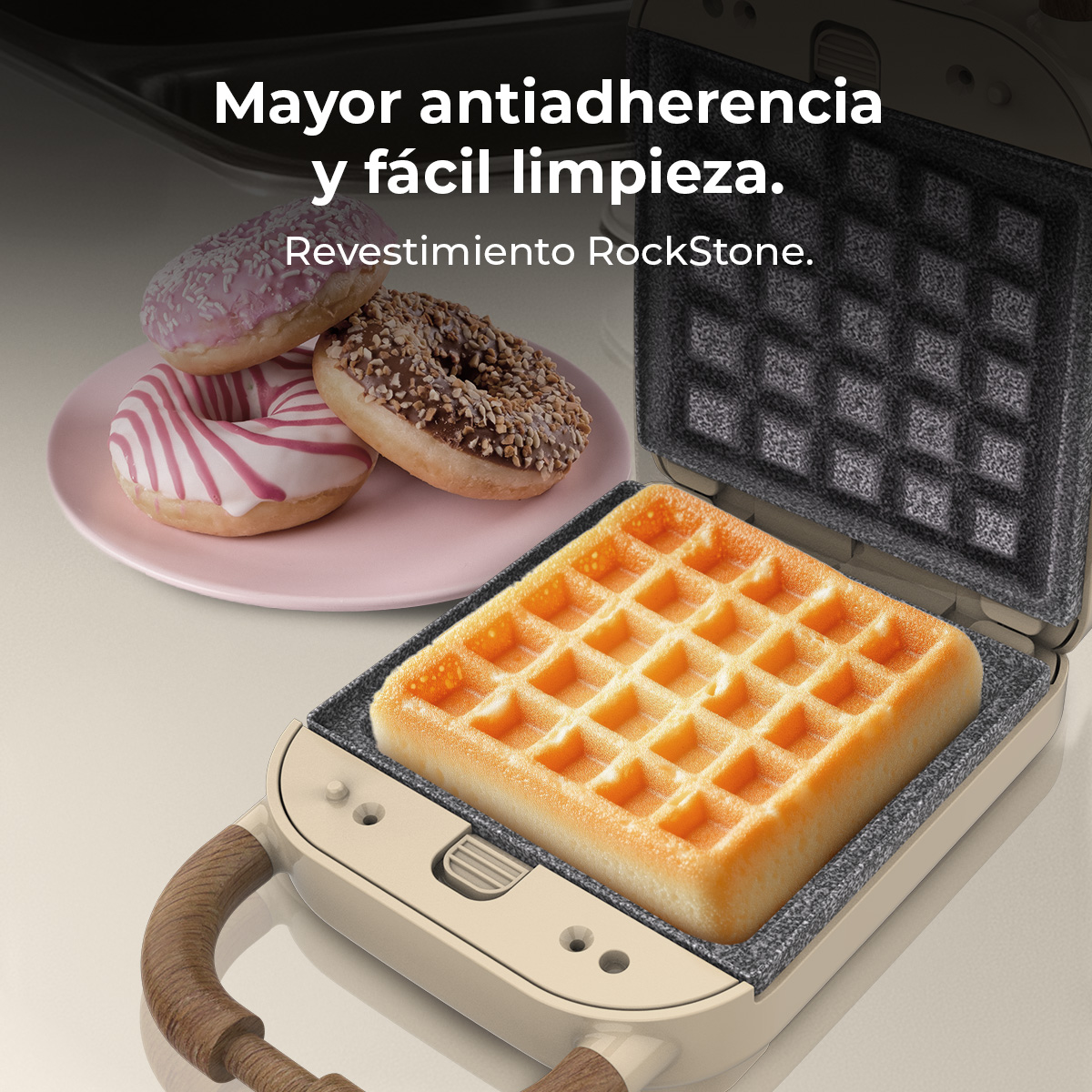 Una macchina per waffle con un waffle cotto all'interno e tre ciambelle su un piatto rosa sullo sfondo.