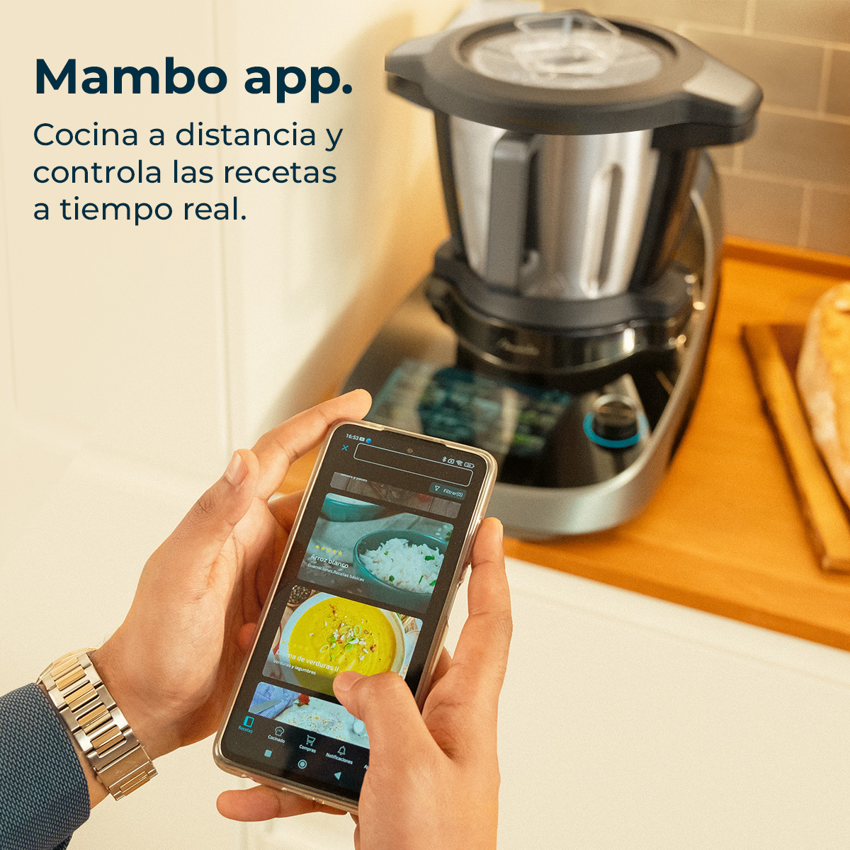 Una persona sostiene un teléfono que muestra la interfaz de una aplicación de cocina, con un procesador de alimentos en el fondo. El texto dice Mambo app.