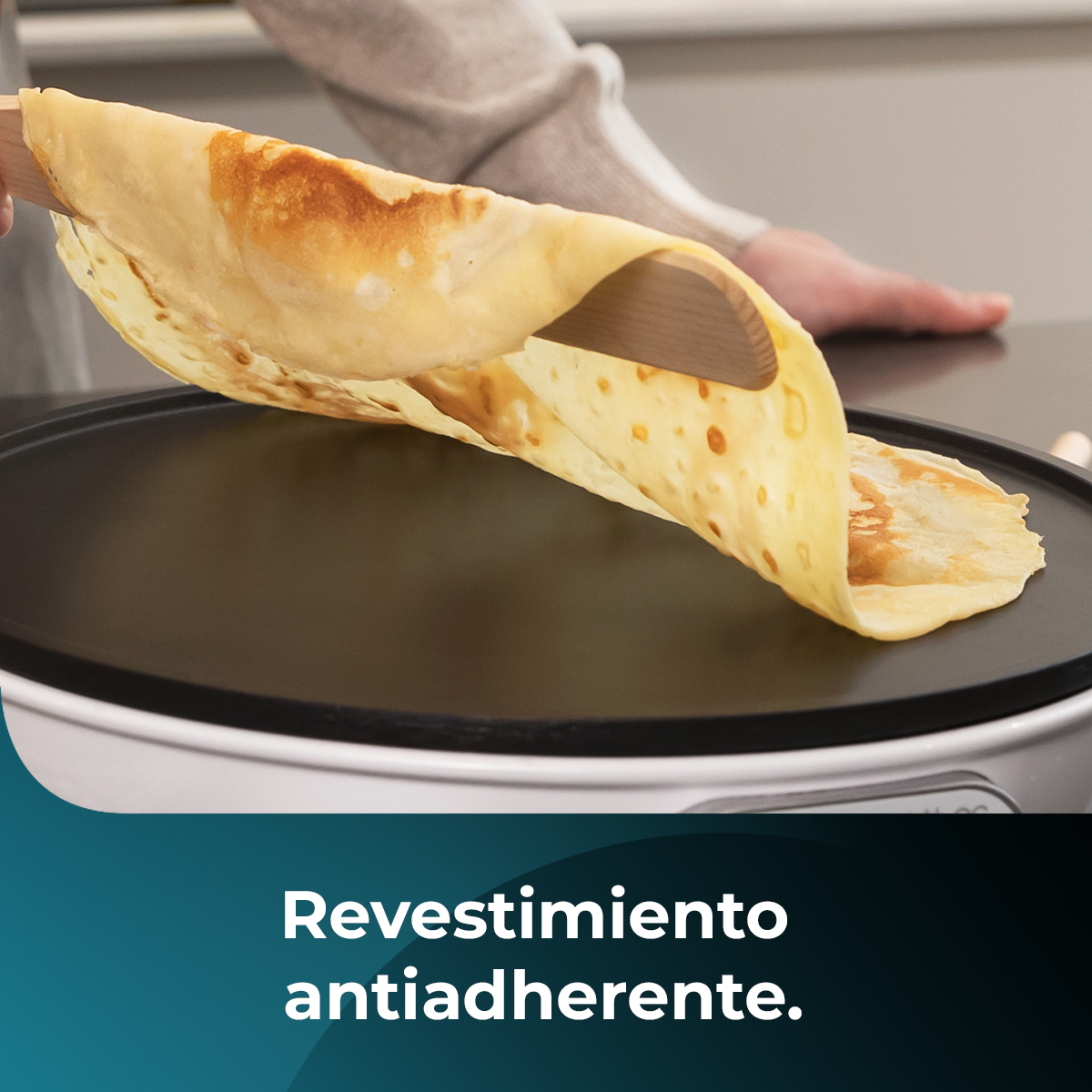 Una persona capovolge una crepe su una superficie di cottura nera con una spatola di legno. La crepe è dorata e leggermente arricciata.