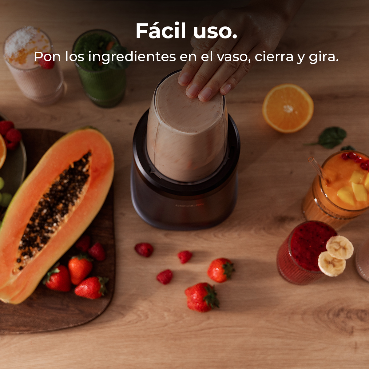 Una mano colocando un vaso de licuadora en su base. Frutas y batidos en una mesa de madera, incluyendo papaya, fresas y naranjas.
