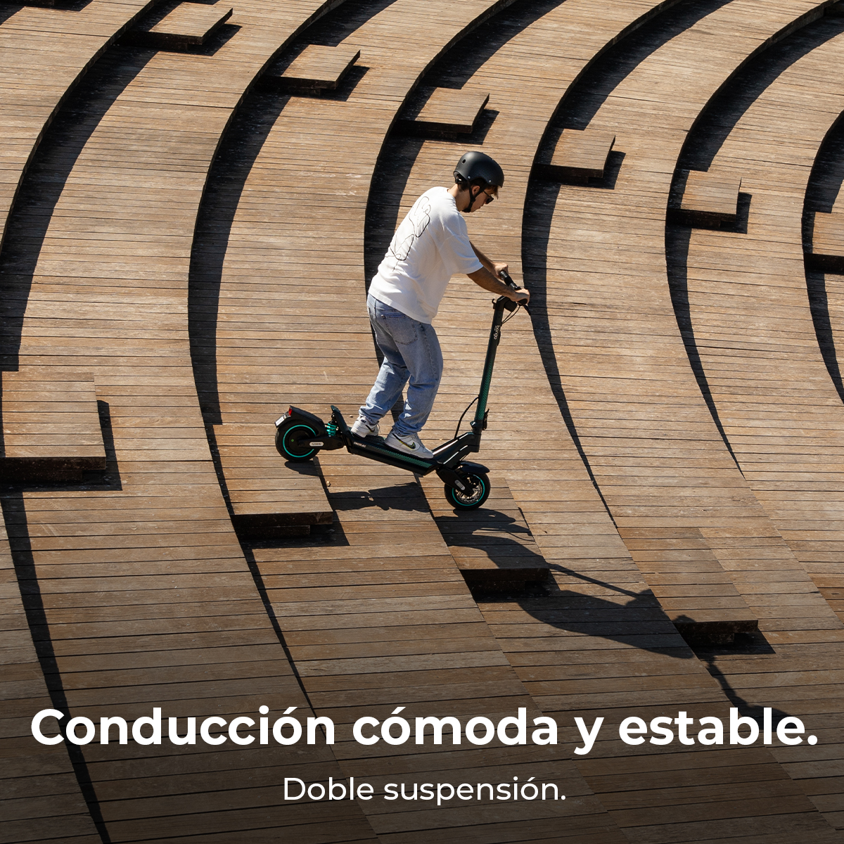 Una persona monta un patinete verde y negro en una rampa de madera. Lleva casco, camisa blanca y vaqueros. El fondo es de madera.