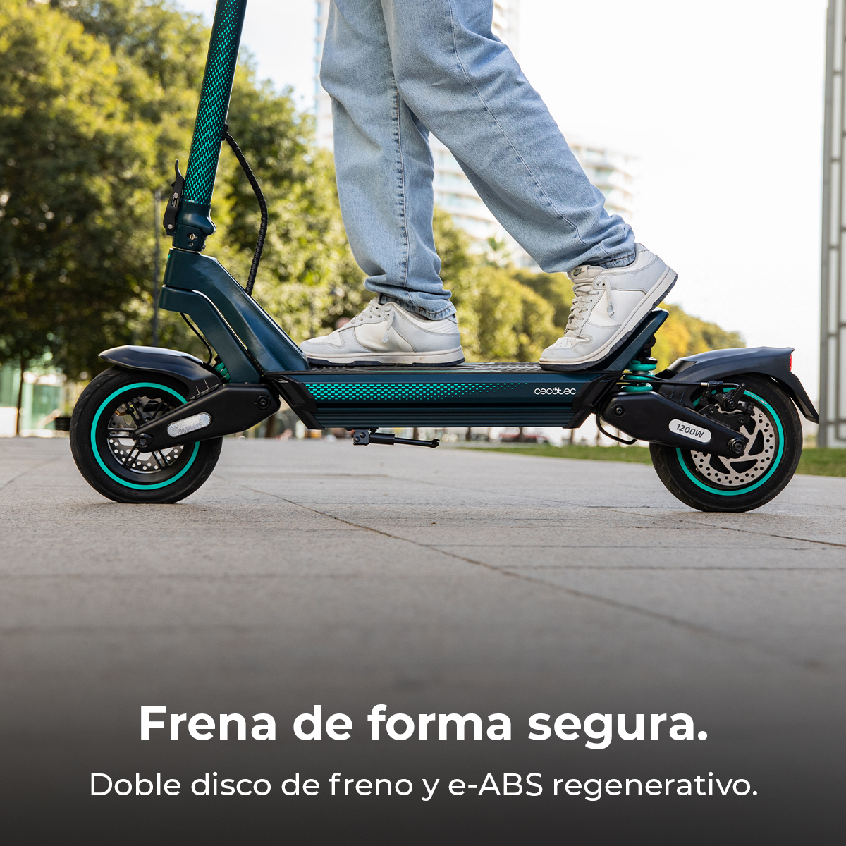Una persona monta un patinete eléctrico verde azulado y negro sobre un camino pavimentado. El patinete tiene detalles verde azulado y se ven zapatos blancos.