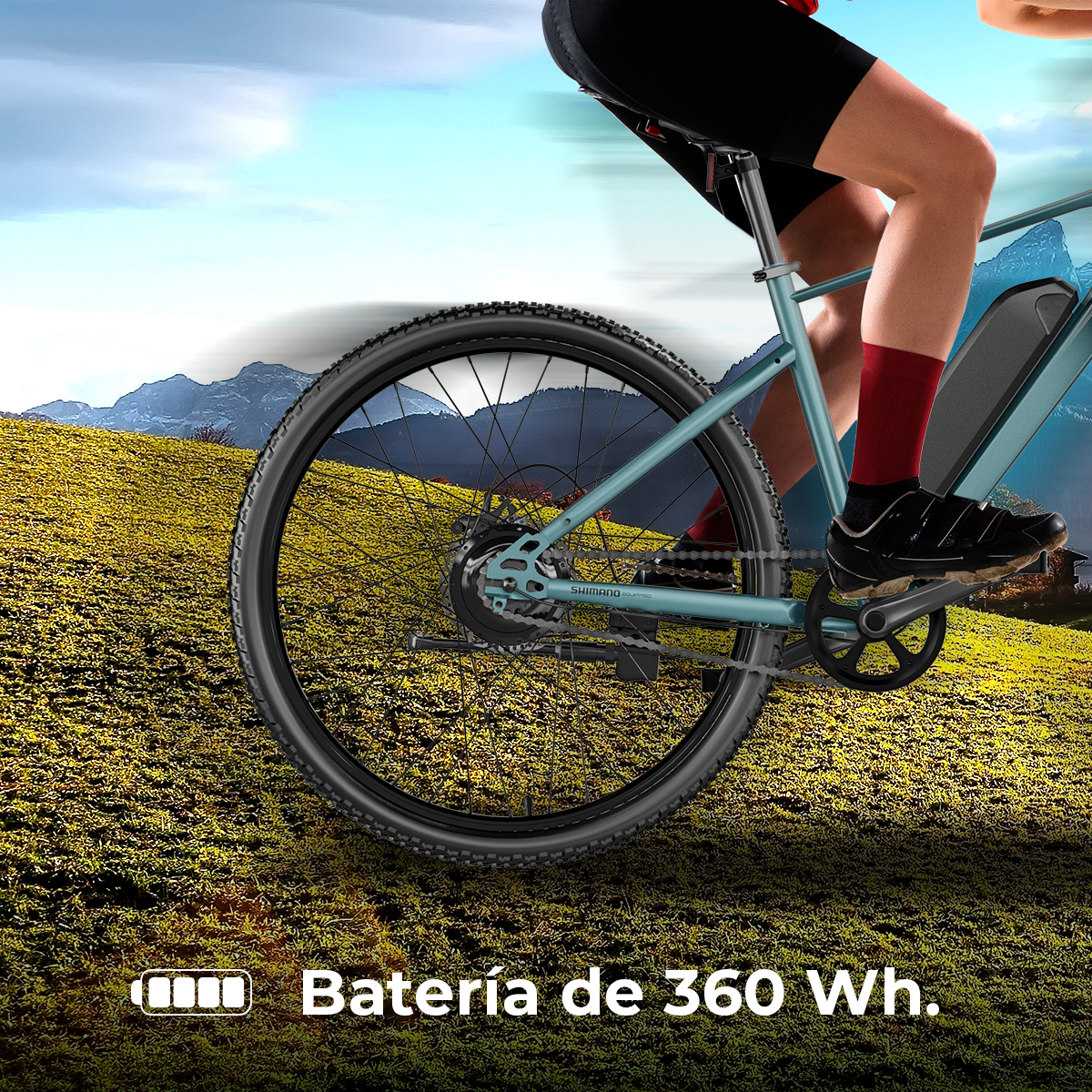 Un ciclista guida una bici blu su una collina erbosa. Montagne e cielo blu sullo sfondo. La bici ha una batteria da 360 Wh.
