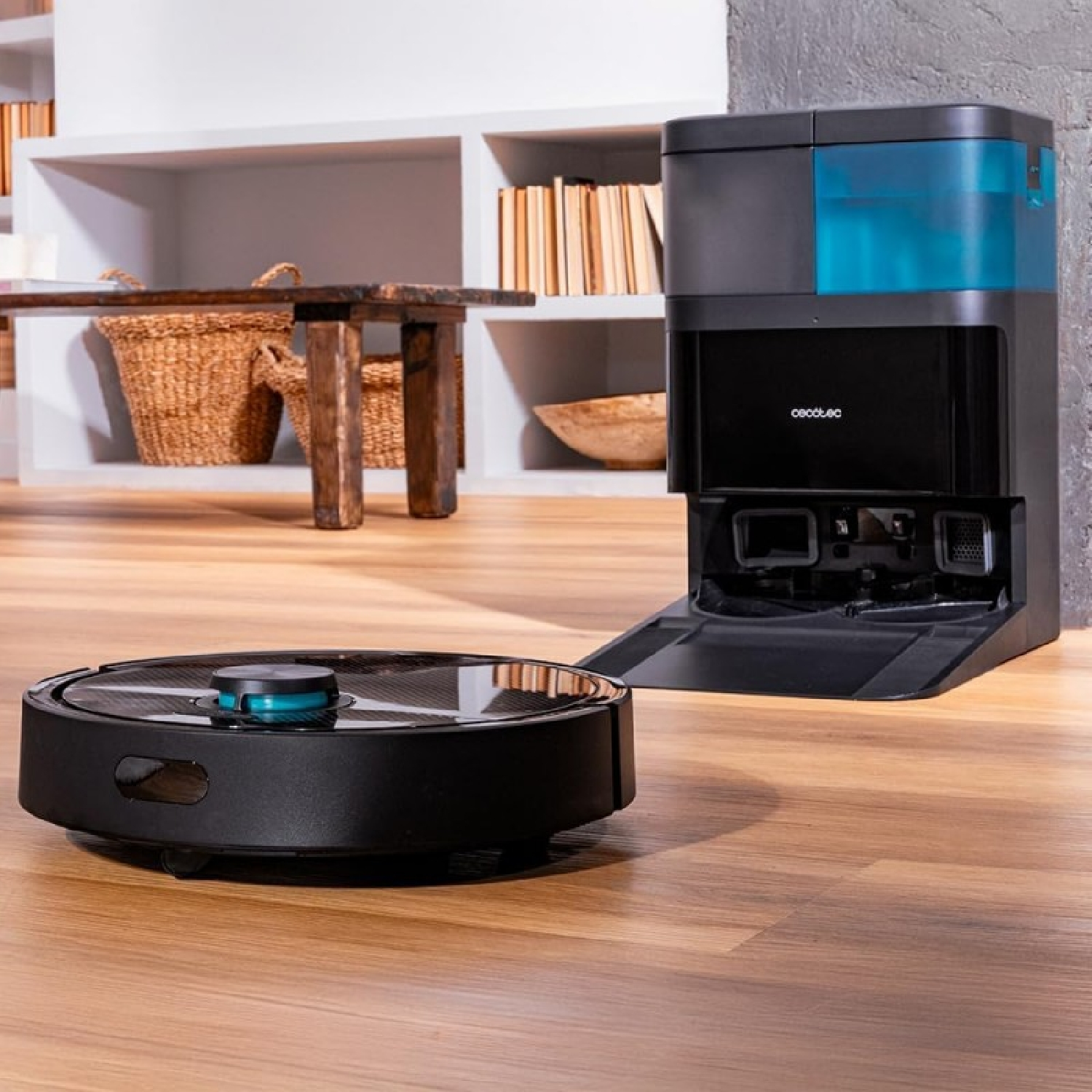 Una aspiradora robot negra está sobre un piso de madera, cerca de una estación de carga. Una mesa y estantes con decoración se ven atrás.