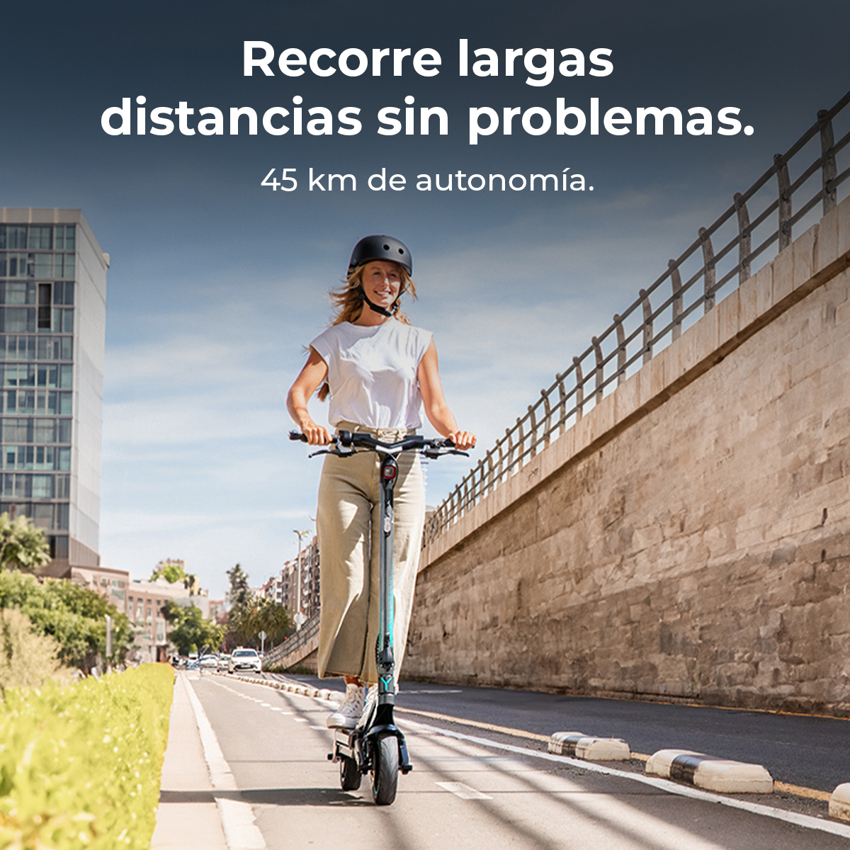 Una mujer monta un patinete eléctrico en una carretera. Lleva casco y ropa informal. Edificios y un puente al fondo.