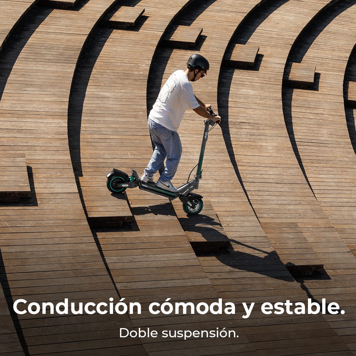 Un hombre monta un patinete eléctrico sobre una estructura de madera. Lleva casco, vaqueros y una camisa blanca. El patinete es gris y negro.