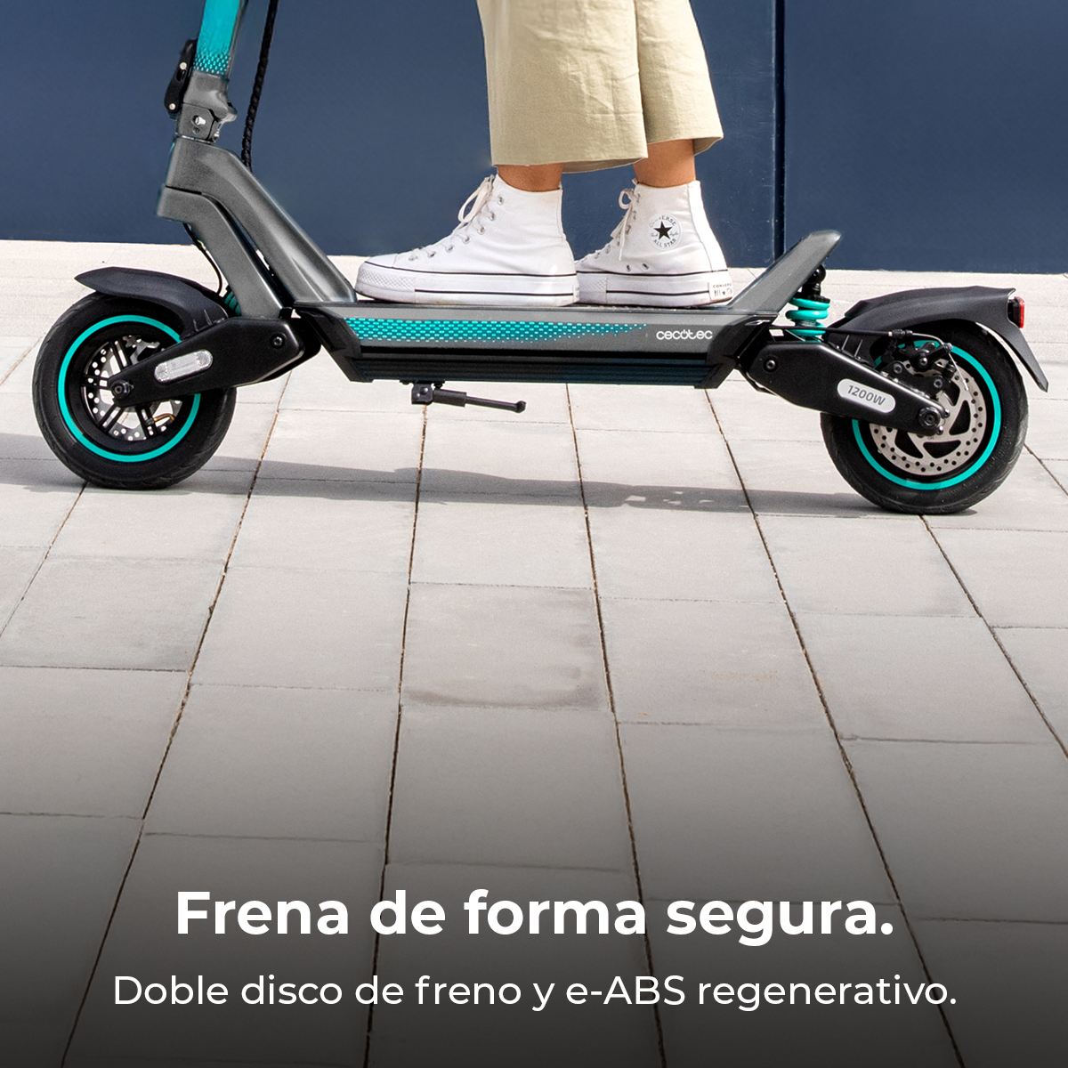 Persona en un patinete eléctrico gris con detalles turquesa, usando zapatillas blancas. El patinete está sobre baldosas.