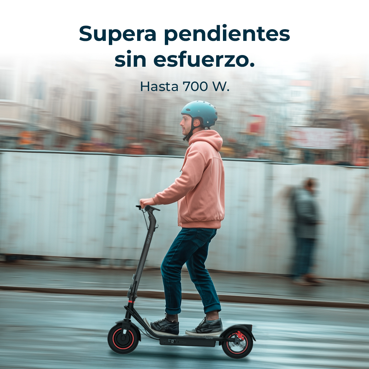 Un uomo guida un monopattino elettrico per strada. Indossa un casco, una felpa con cappuccio rosa e jeans. Lo sfondo è sfocato.