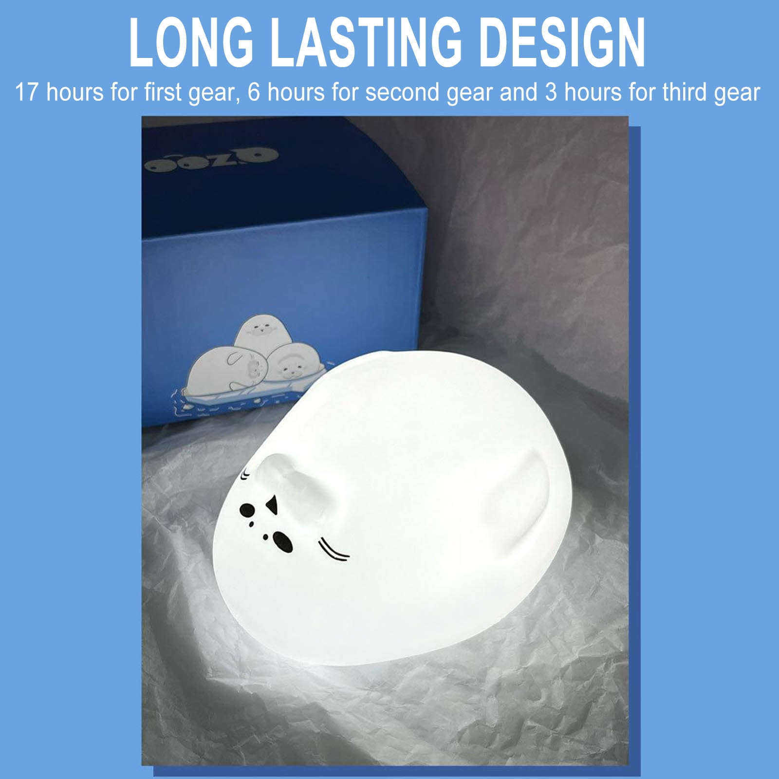 Eine weiße, seehundförmige Lampe liegt auf Papier. Dahinter befindet sich eine blaue Box mit Seehunddesign.