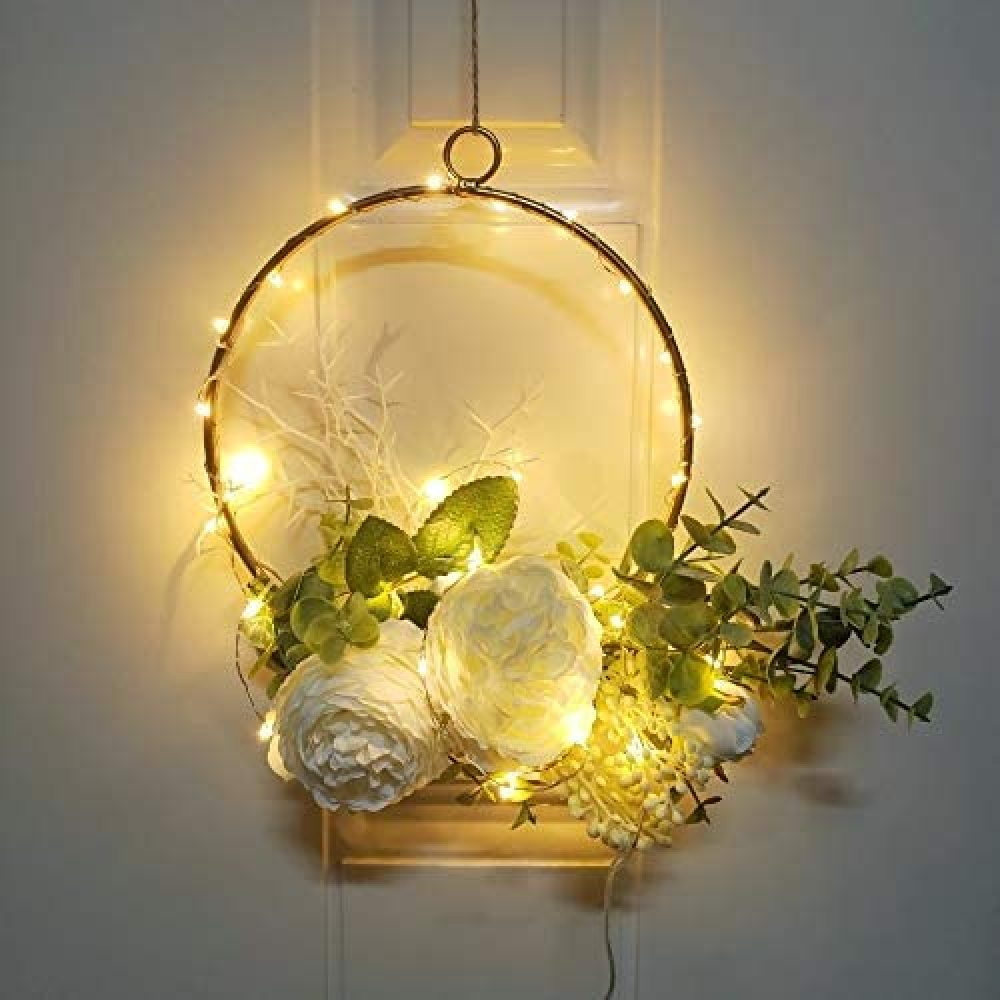 Ein dekorativer Kranz mit goldenem Ring, Lichterketten, weißen Blumen und grünen Blättern. Er hängt an einer weißen Tür.