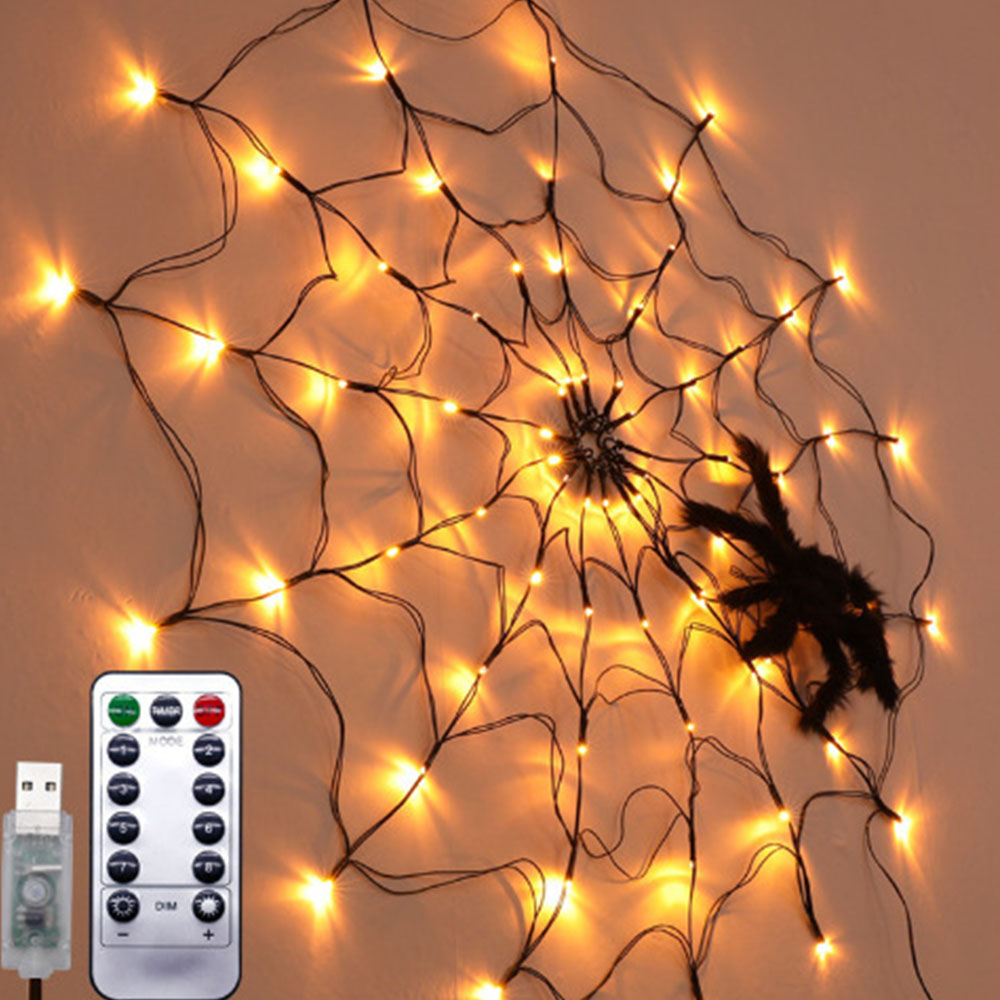 Eine Spinnennetzdekoration mit warmen Lichtern, einer schwarzen Spinne und einer Fernbedienung. USB-Kabel sichtbar.