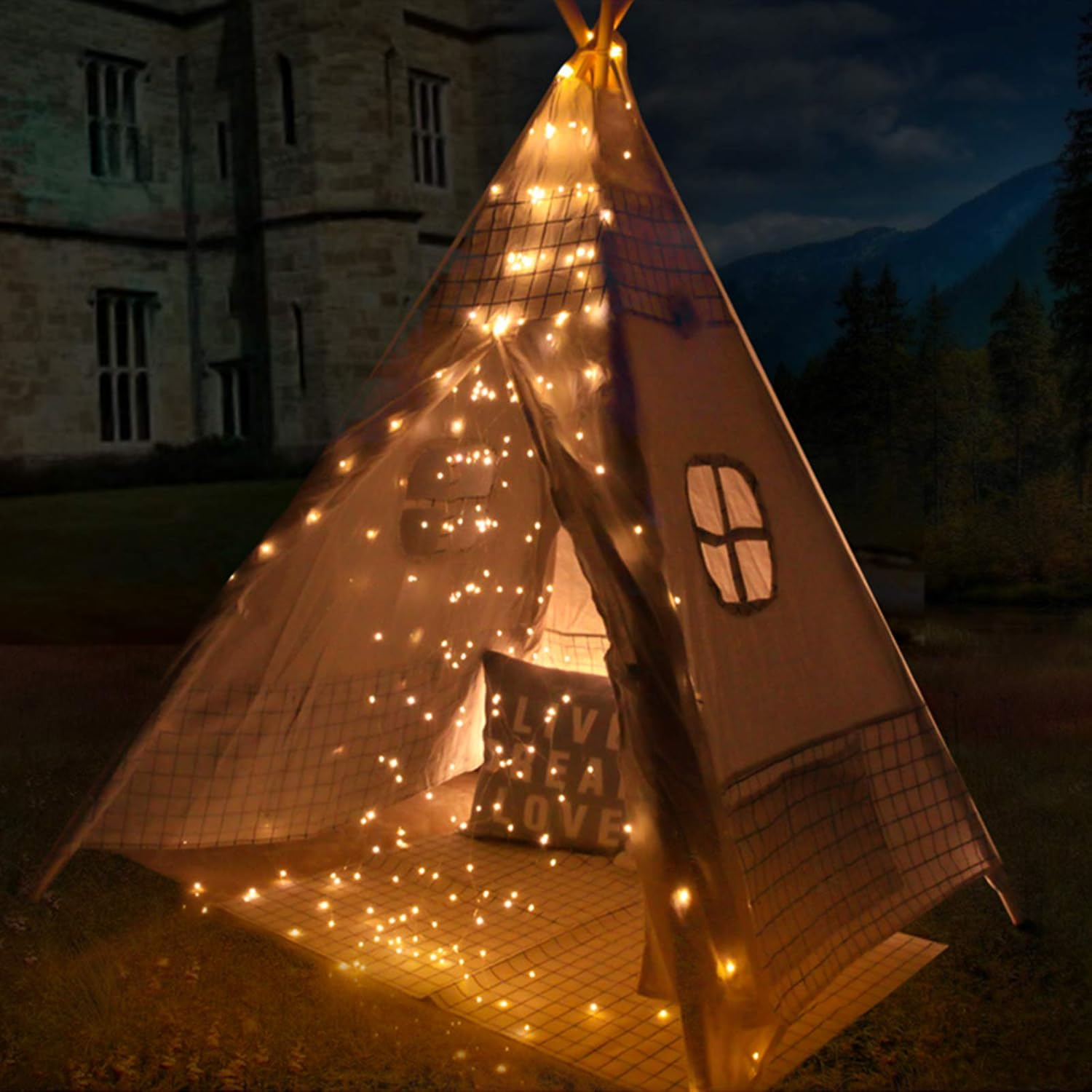 Ein beleuchtetes Tipi-Zelt mit Lichterketten steht auf Gras. Im Hintergrund ist ein Gebäude zu sehen. Ein Kissen darin liest 'lebe, lache, liebe'.