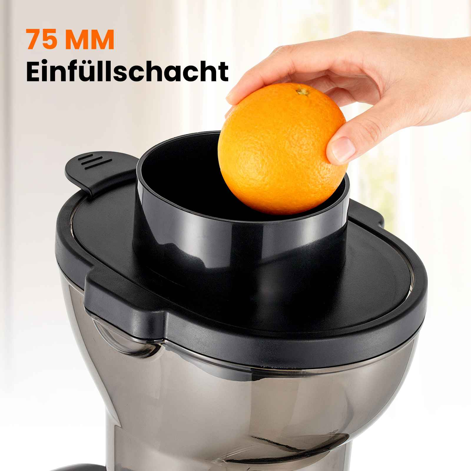Eine Hand legt eine Orange in den schwarzen Trichter eines Entsafters. Die Maschine hat schwarze und transparente Teile.