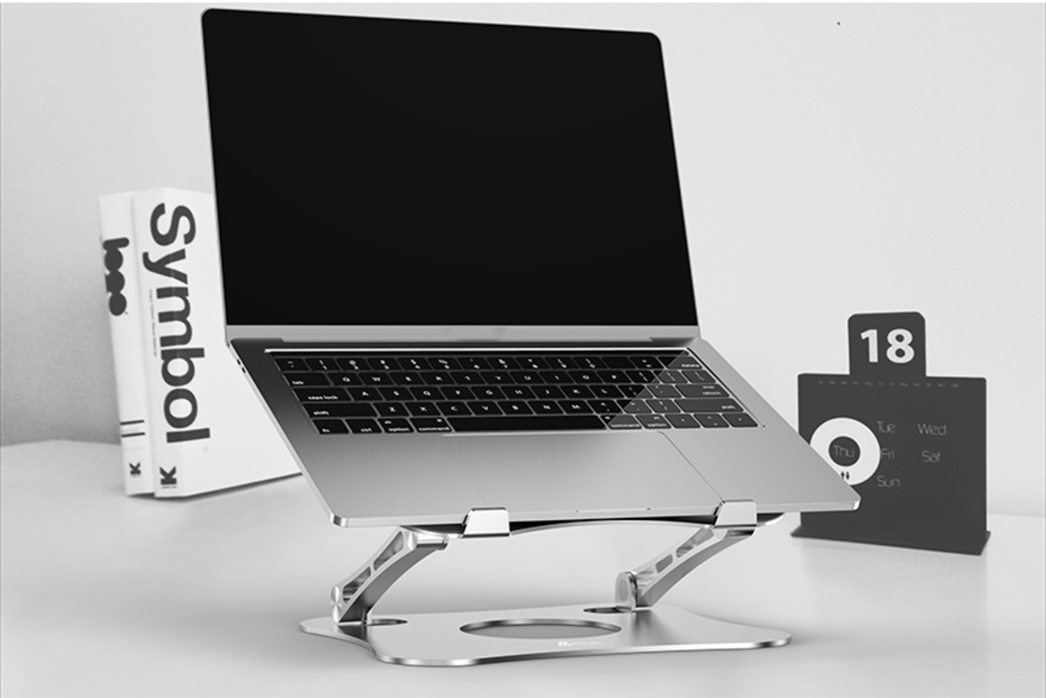 Laptop na srebrnym stojaku, czarny ekran, widoczna klawiatura. Książki i kalendarz w tle. Data to 18.