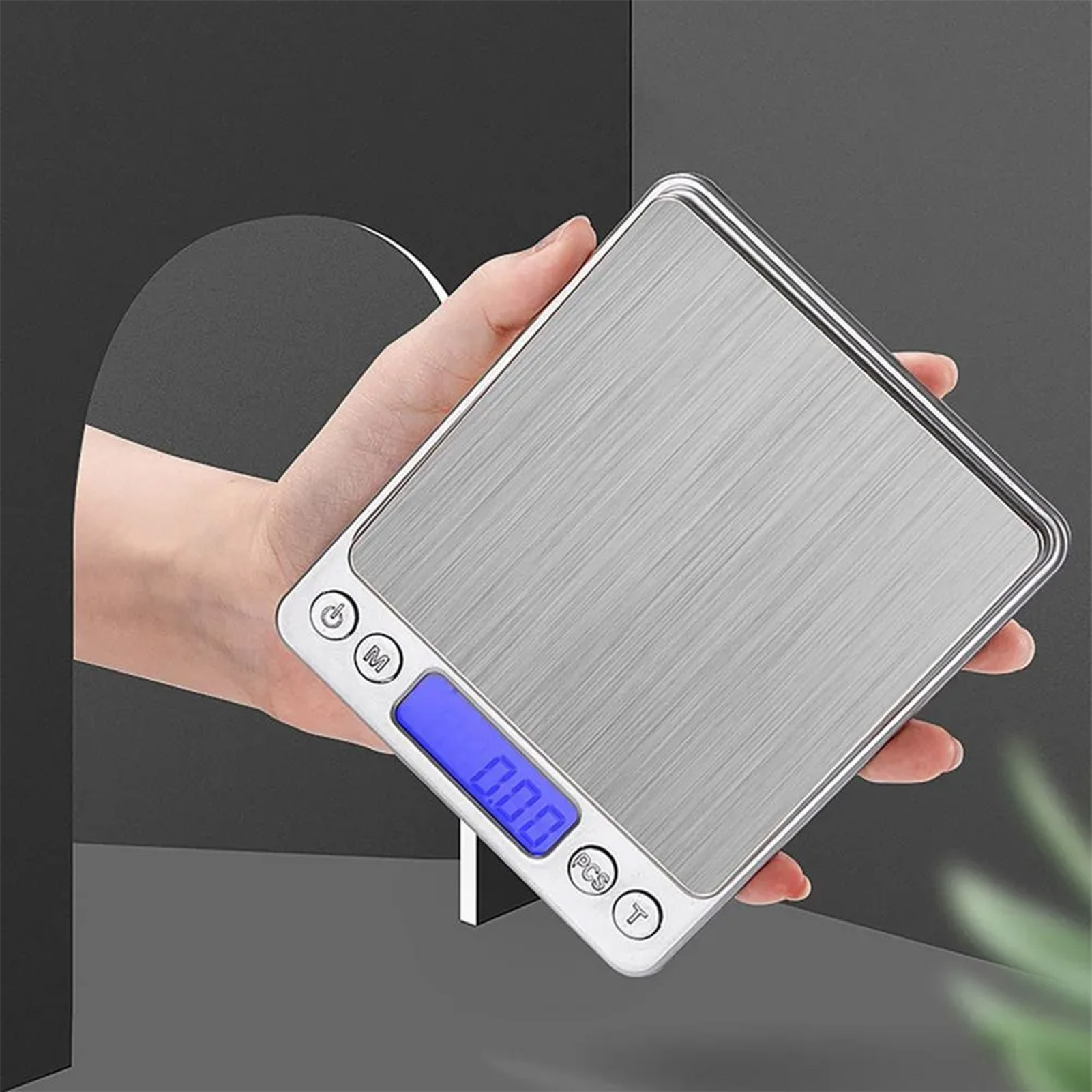Eine Hand hält eine silberne Digitalwaage. Das Display zeigt 0,00 an. Knöpfe sind auf der Waage sichtbar. Ein schwarzer und grauer Hintergrund.