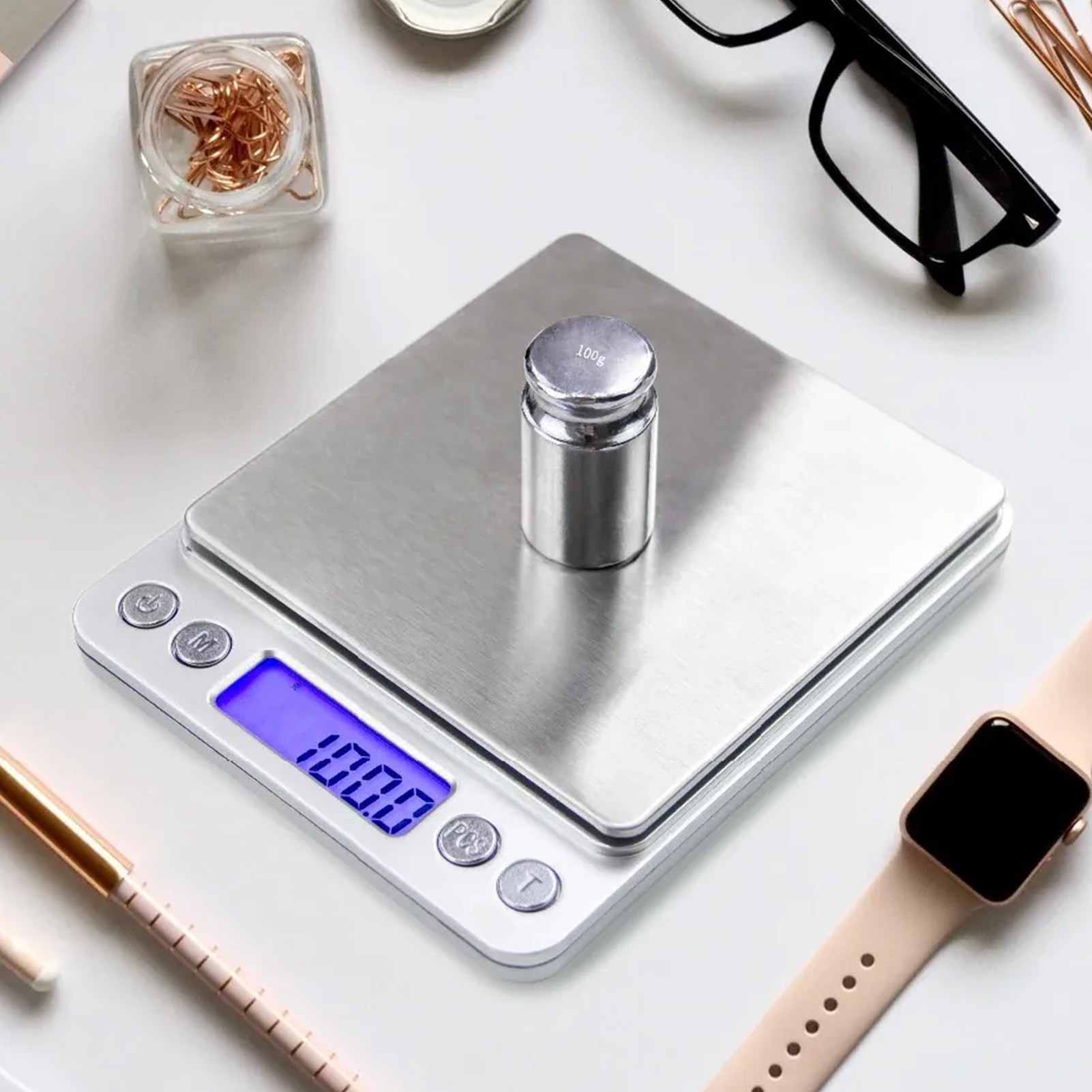 Eine Digitalwaage mit einem 100g-Gewicht. Eine Uhr, Brille und Bürobedarf umgeben die Waage auf weißer Oberfläche.