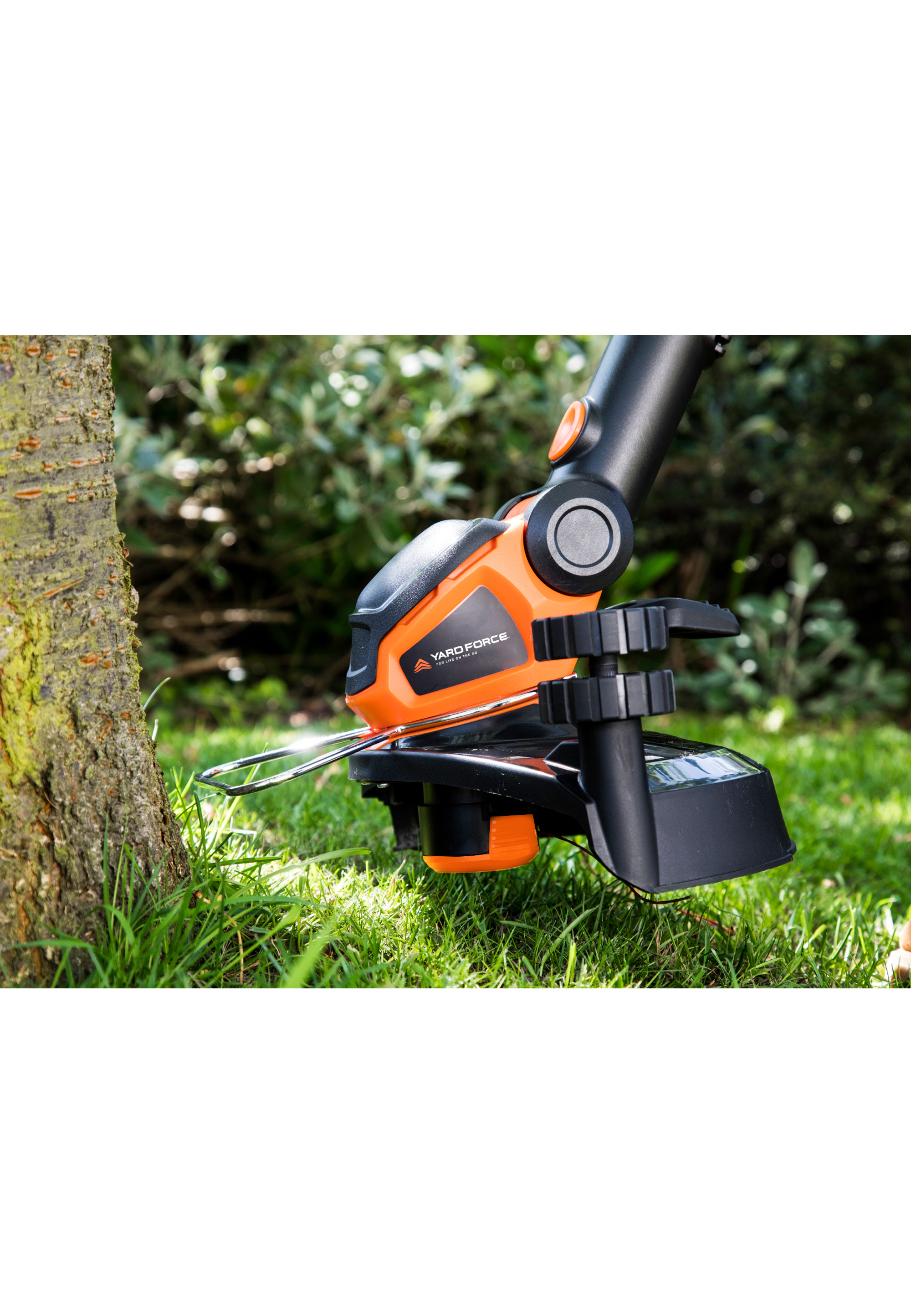 Ein orangefarbener und schwarzer Yard Force-Rasentrimmer schneidet Gras in der Nähe eines Baumes. Grünes Laub ist im Hintergrund.