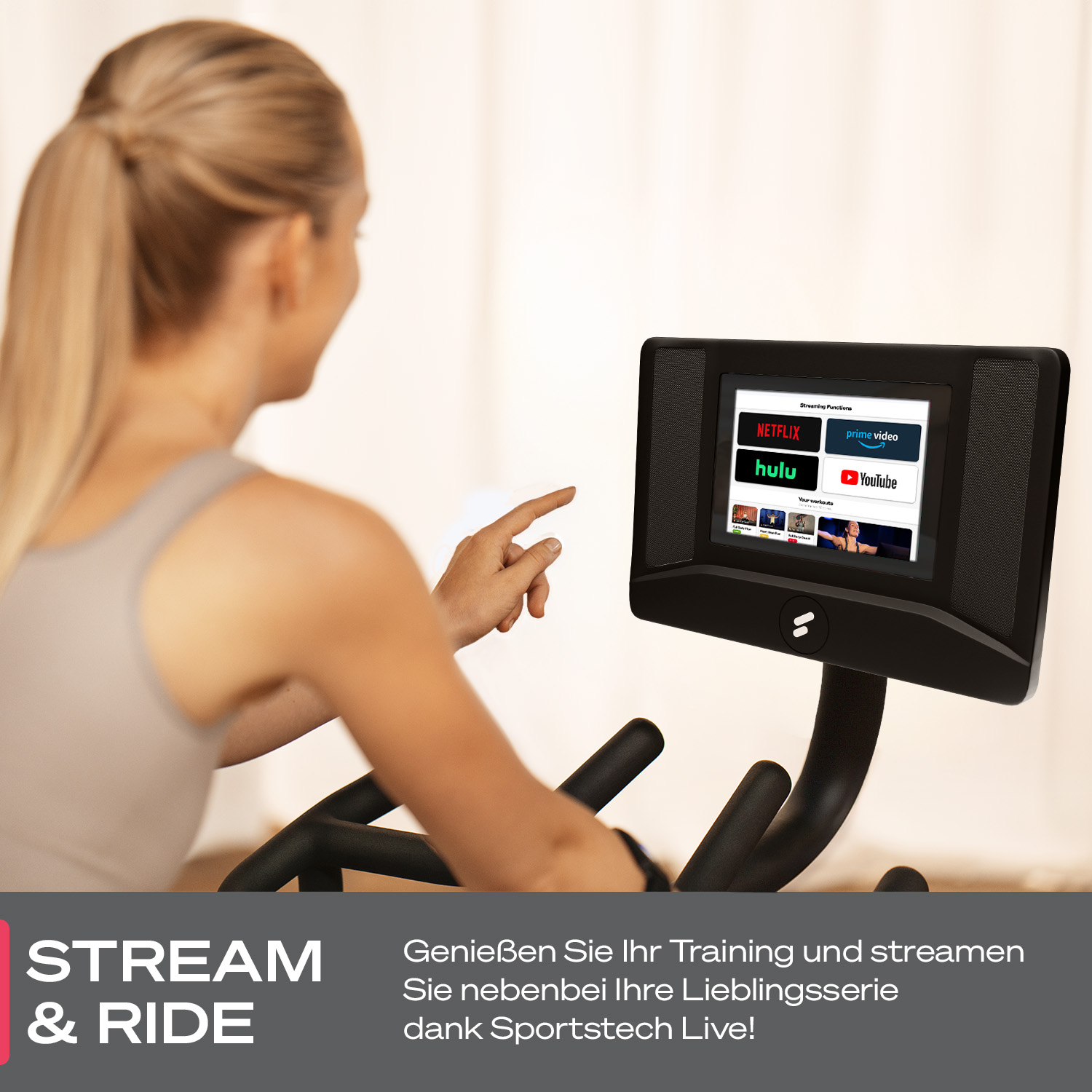 Eine Frau benutzt ein Trainingsgerät mit einem Bildschirm, auf dem Streaming-Dienste angezeigt werden. Sie zeigt auf den Bildschirm. Der Hintergrund ist unscharf.