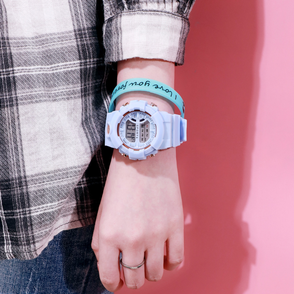 Ein Handgelenk mit einer blauen Uhr und einem Armband. Die Person trägt ein kariertes Hemd und einen Silberring. Der Hintergrund ist rosa.