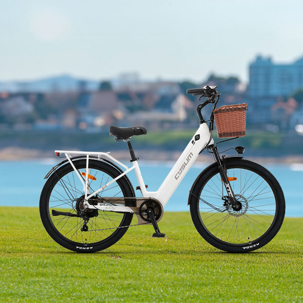 Biały rower elektryczny z brązowym koszem na zielonej trawie w pobliżu wody i budynków w tle.
