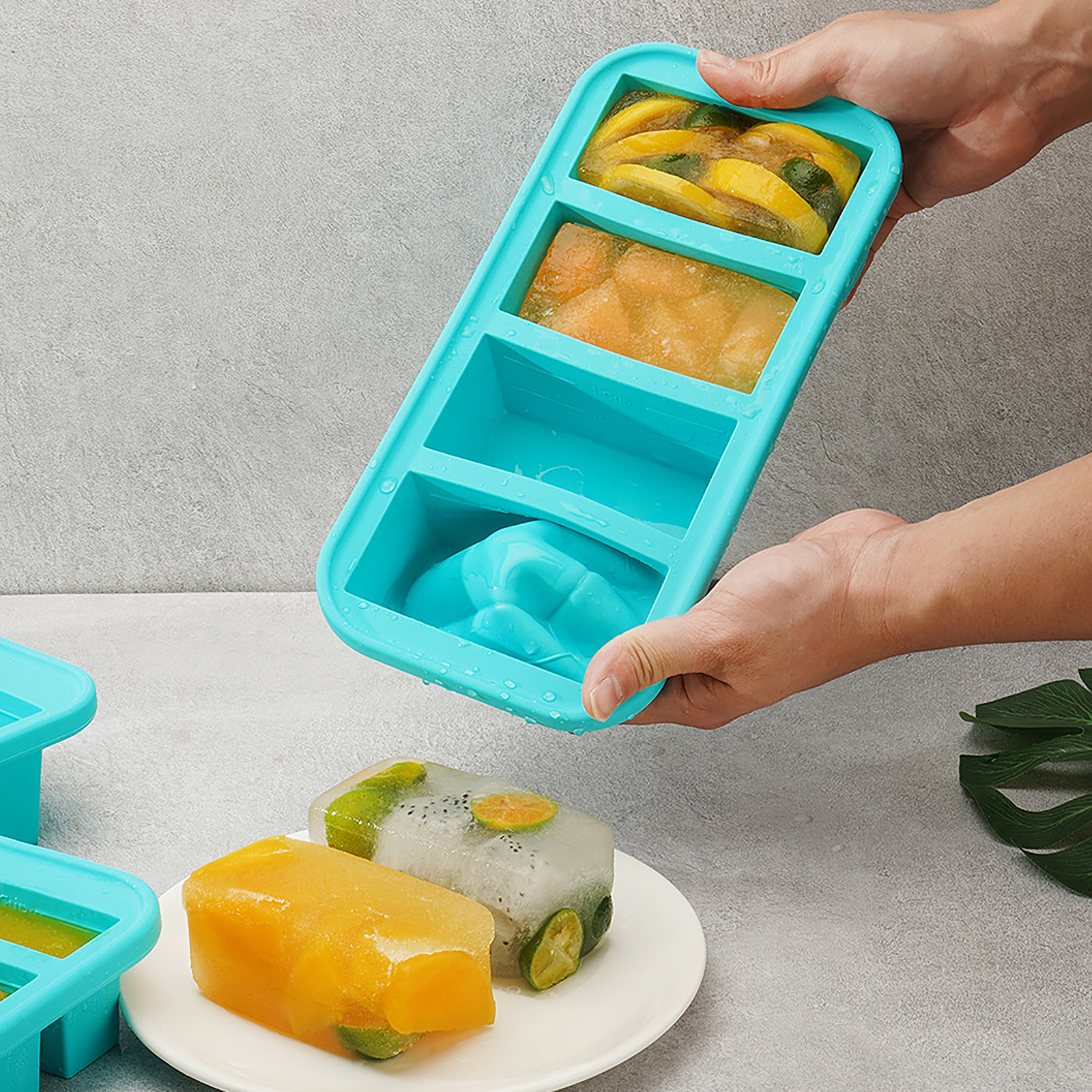 Eine Hand hält eine blaue Eiswürfelschale mit Obst darin. Gefrorene Würfel liegen auf einem Teller. Grauer Hintergrund.