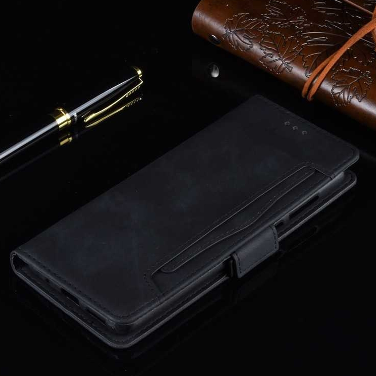 Eine schwarze Leder-Handyhülle mit einem Stift und einem braunen Leder-Notizbuch auf schwarzer, reflektierender Oberfläche.