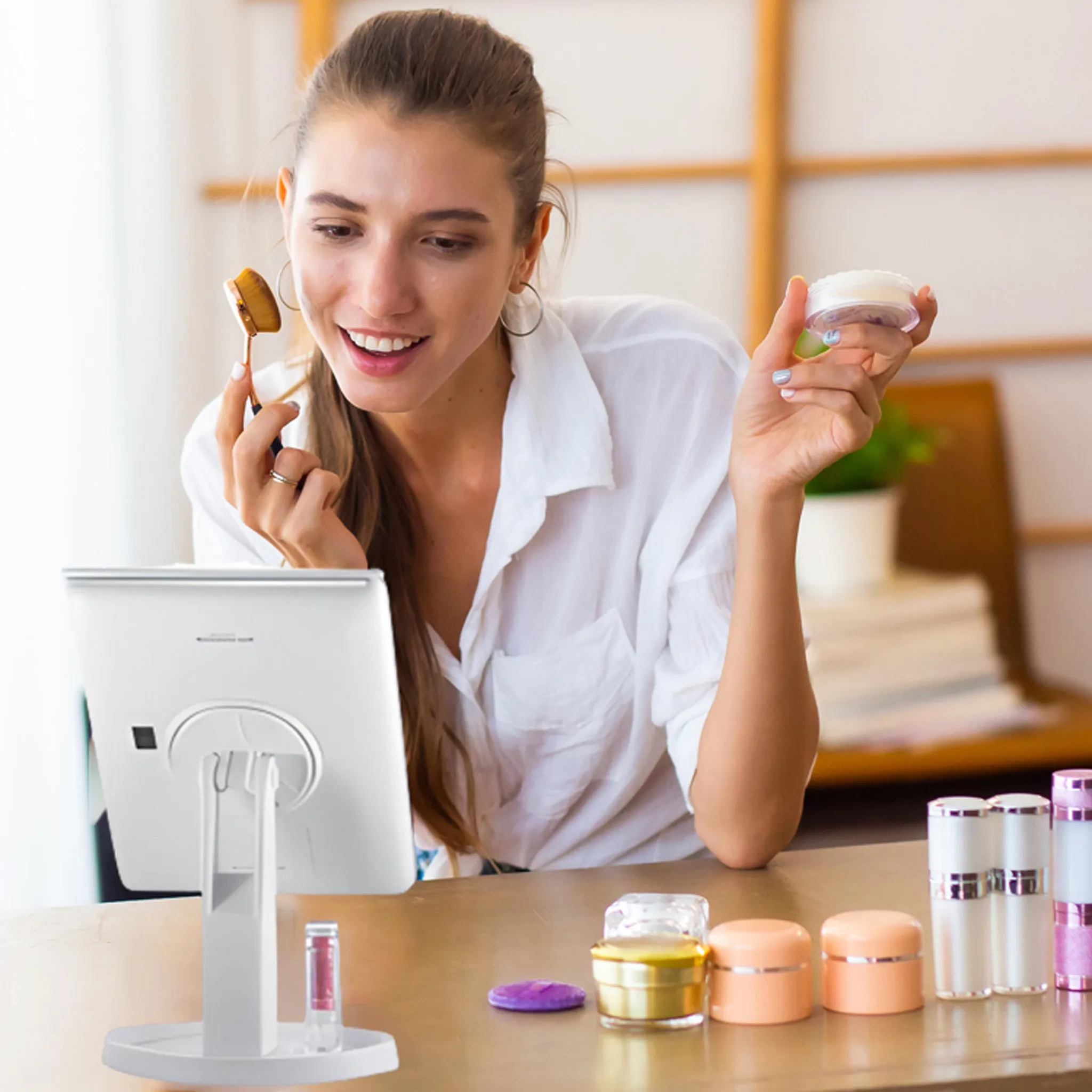 Frau mit Pinsel und Creme, die in den Spiegel lächelt. Kosmetik auf dem Tisch. Weißes Hemd, Ohrringe, Make-up.