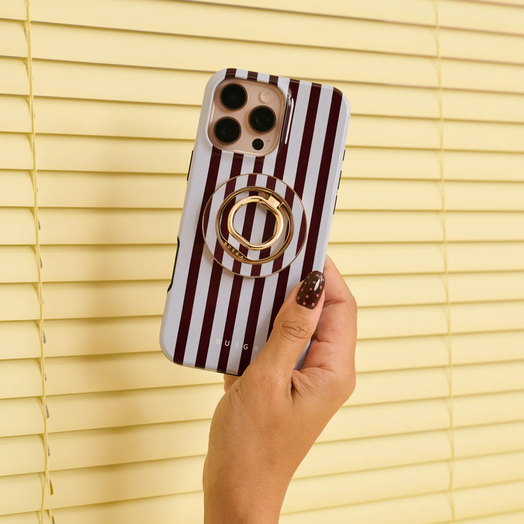 Een hand houdt een telefoon met een gestreepte hoes en een ringhouder vast tegen gele jaloezieën.