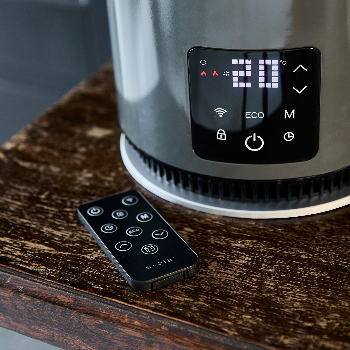 Un riscaldatore grigio scuro con display digitale che mostra 20 gradi. Un telecomando nero su legno.