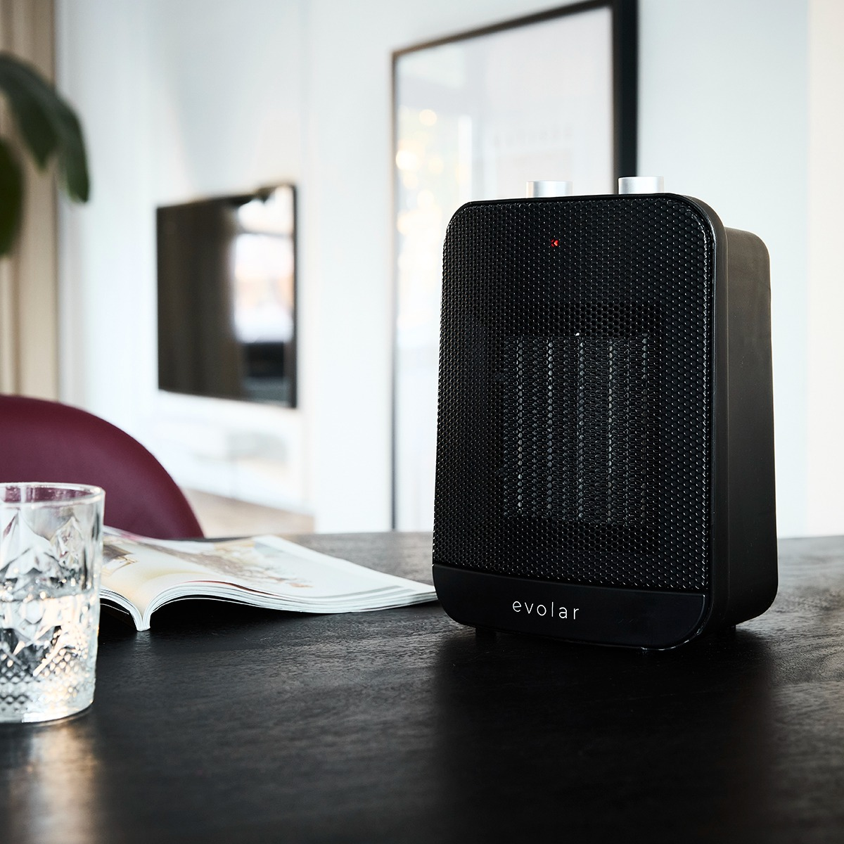 Un termoventilatore Evolar nero è su un tavolo nero. Un bicchiere, una rivista aperta e una TV sono visibili nella stanza.