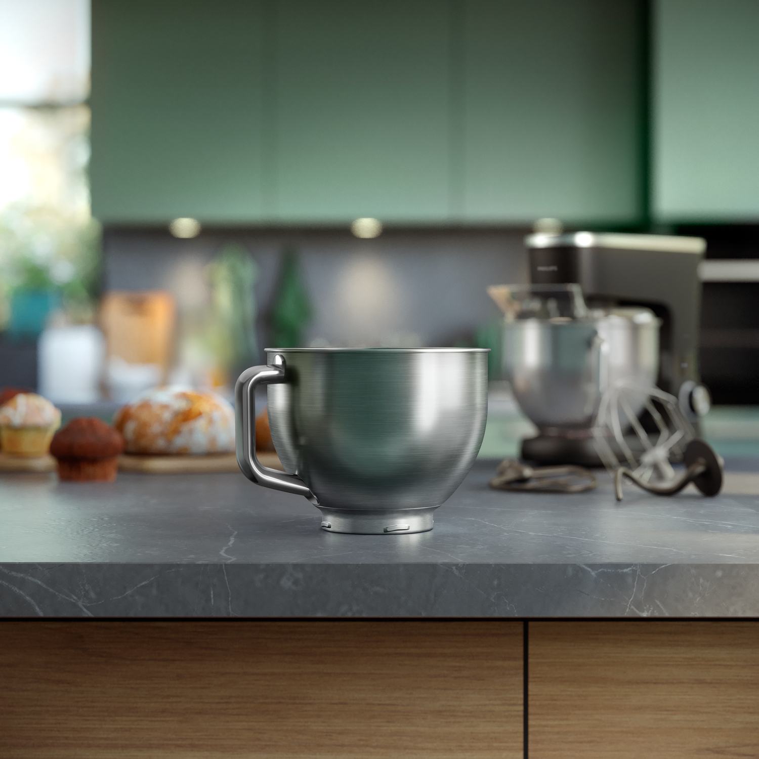 A metal mixing bowl on a countertop. Kitchen appliances, bread, and muffins are in the background. Green cabinets are visible.