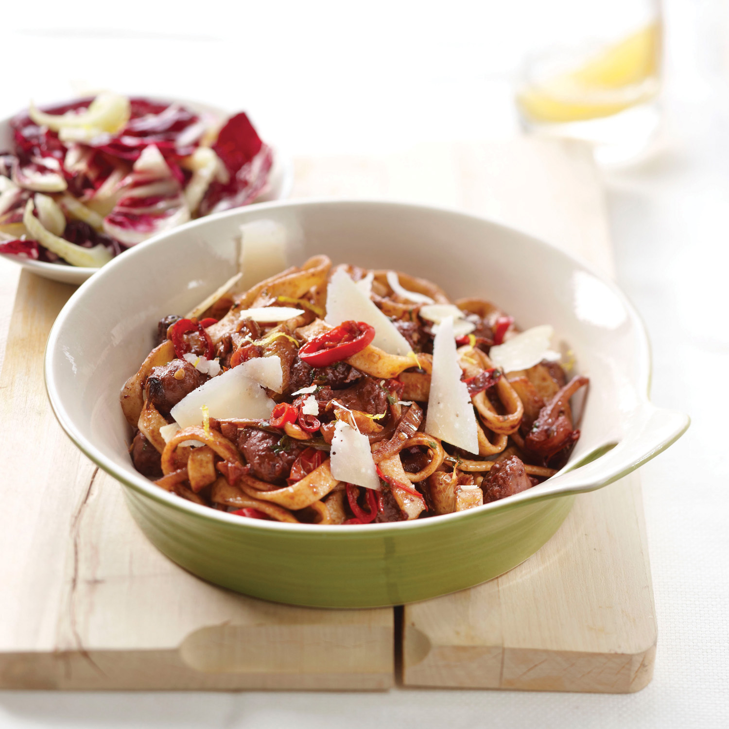 Nudelgericht mit Parmesan, roten Paprika und Wurst in einer grünen Schüssel. Salat in einer weißen Schüssel im Hintergrund. Glas Flüssigkeit.