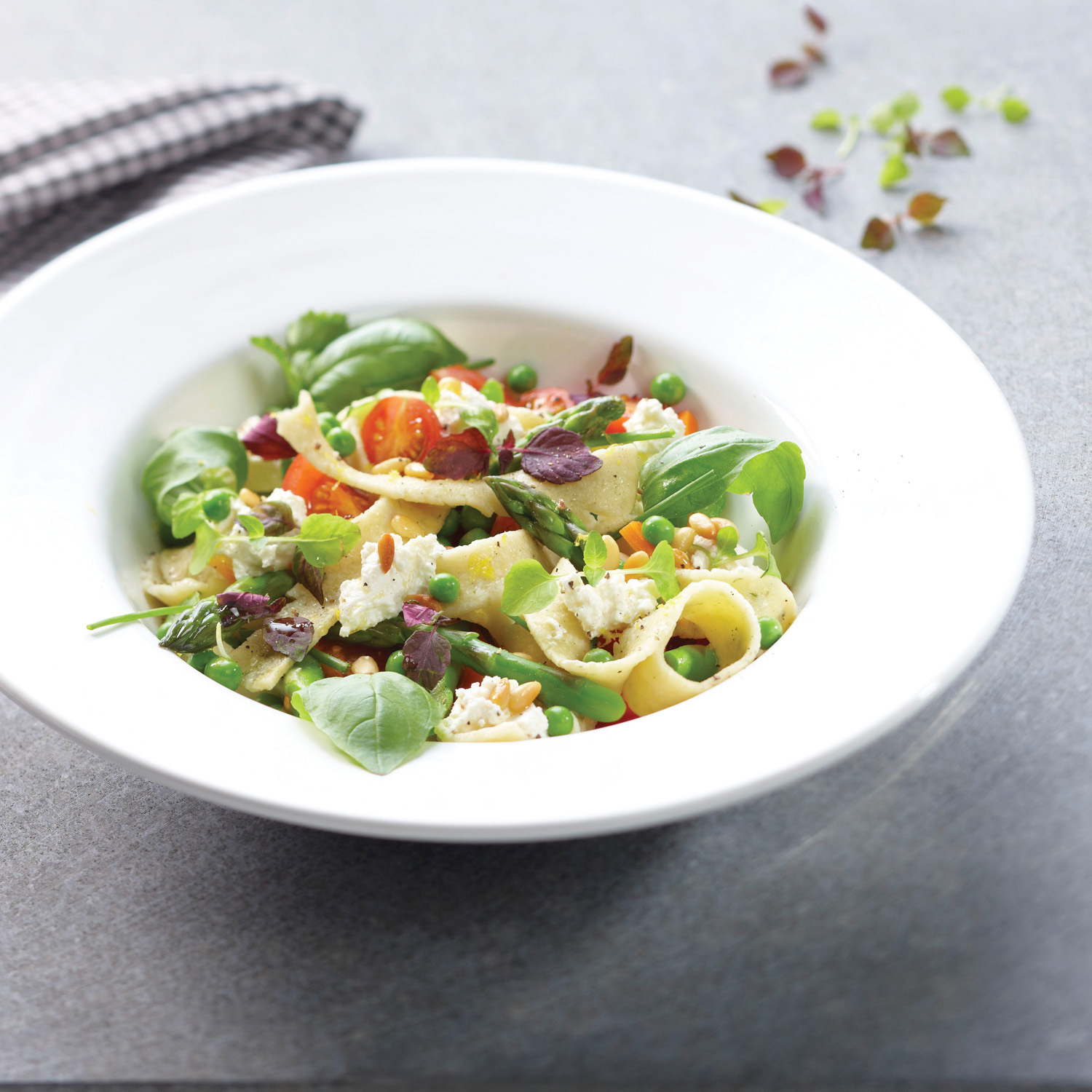 Nudelsalat in einer weißen Schüssel. Er enthält grüne Erbsen, Tomaten, Spargel, Basilikum und Pinienkerne. Der Hintergrund ist eine graue Oberfläche.