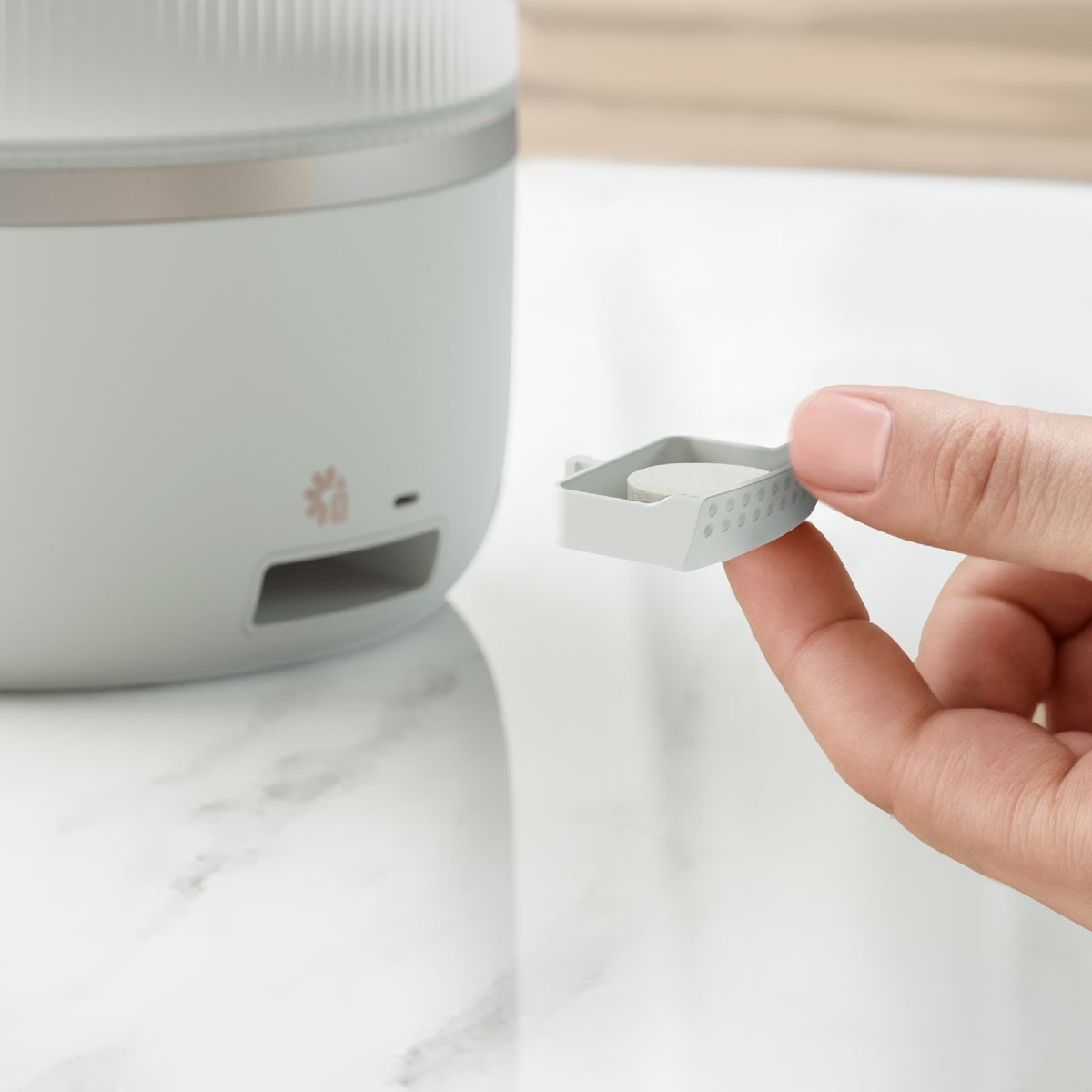 Dłoń trzyma małą, białą tabletkę w szarej tacce. Jasnoszare urządzenie znajduje się w tle, na marmurowej powierzchni.