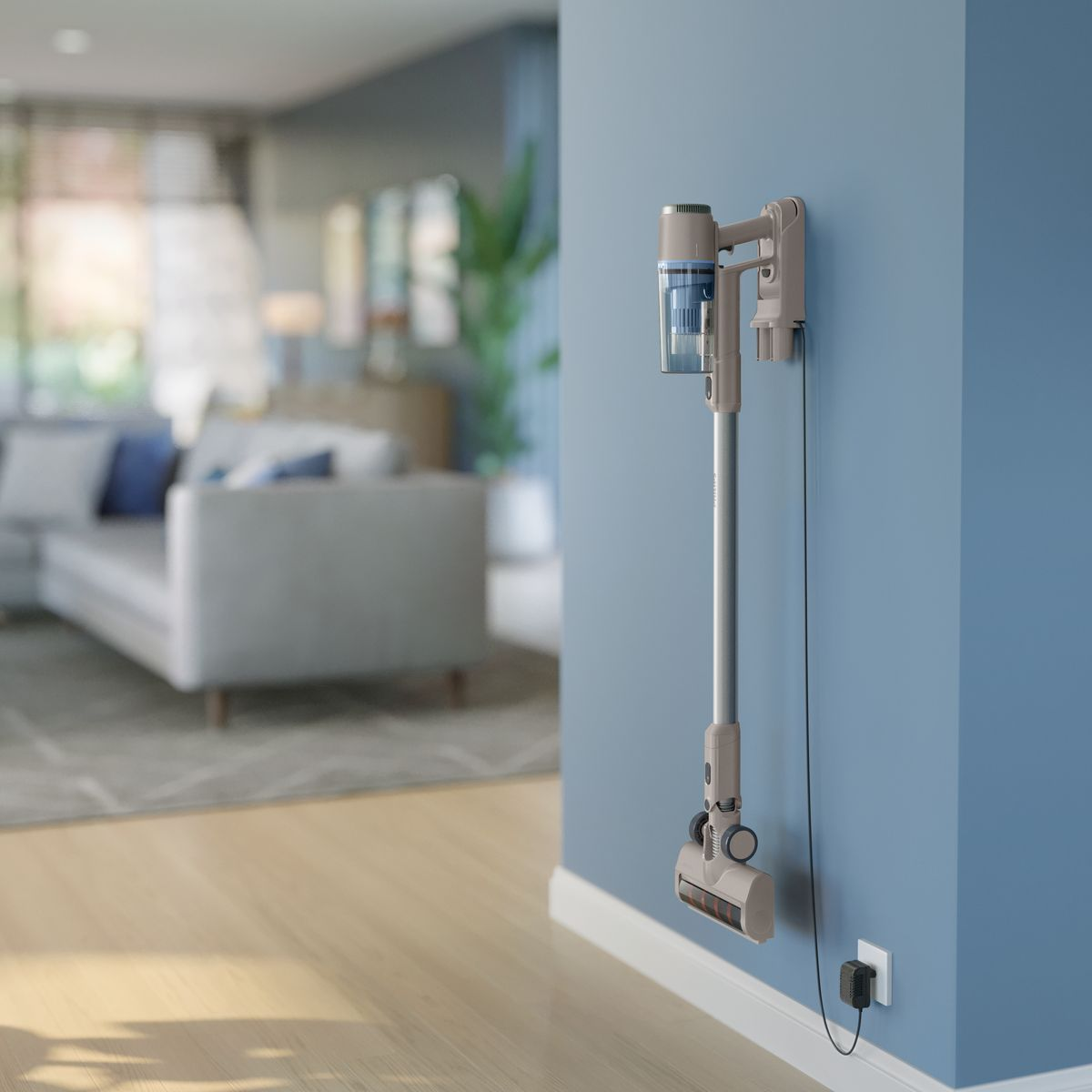 Odkurzacz bezprzewodowy jest zamontowany na niebieskiej ścianie. Odkurzacz jest beżowy i szary. W tle widać salon.