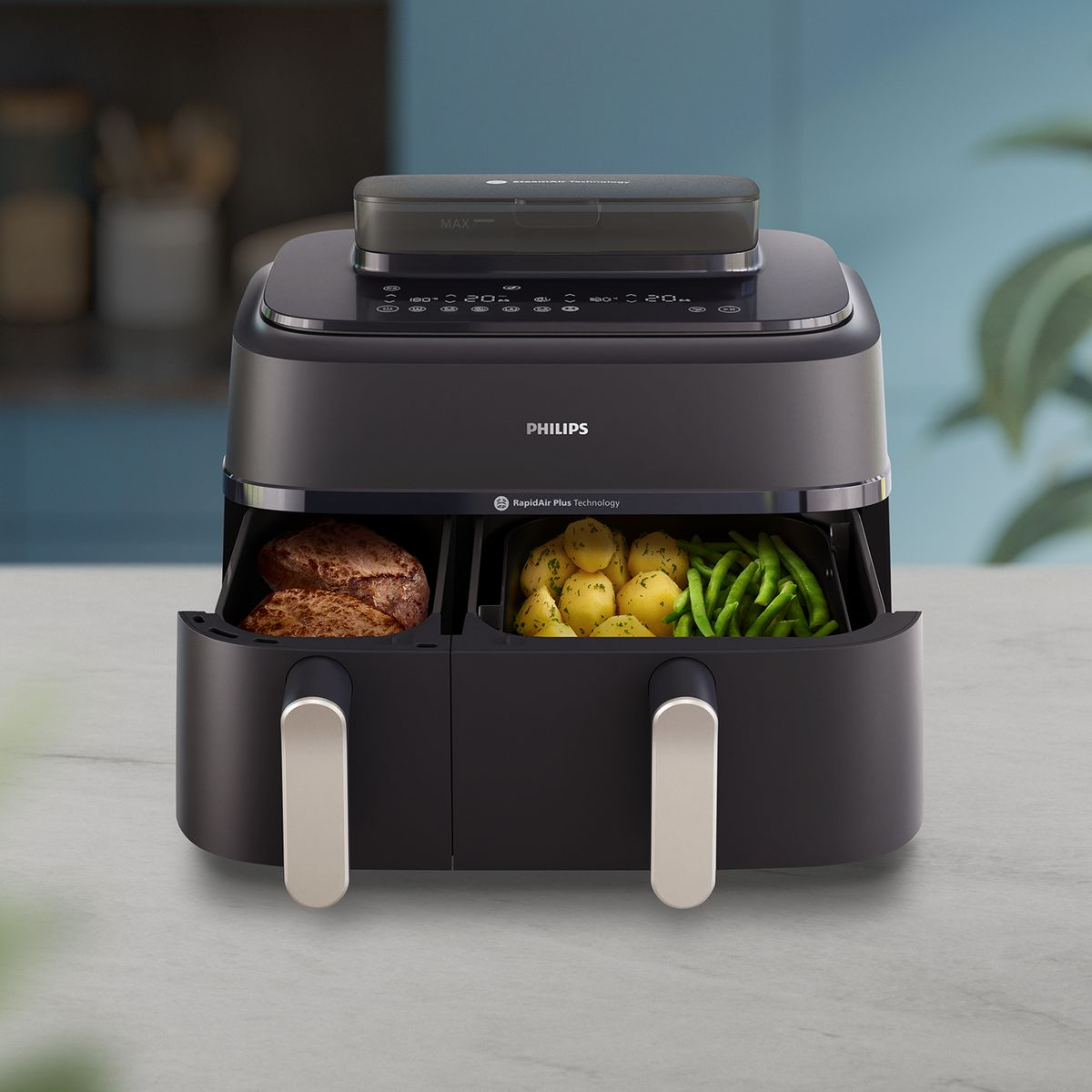 Zwarte Philips heteluchtfriteuse met voedselcompartimenten. Eén heeft vlees, de andere aardappelen en bonen. Op een marmeren oppervlak.