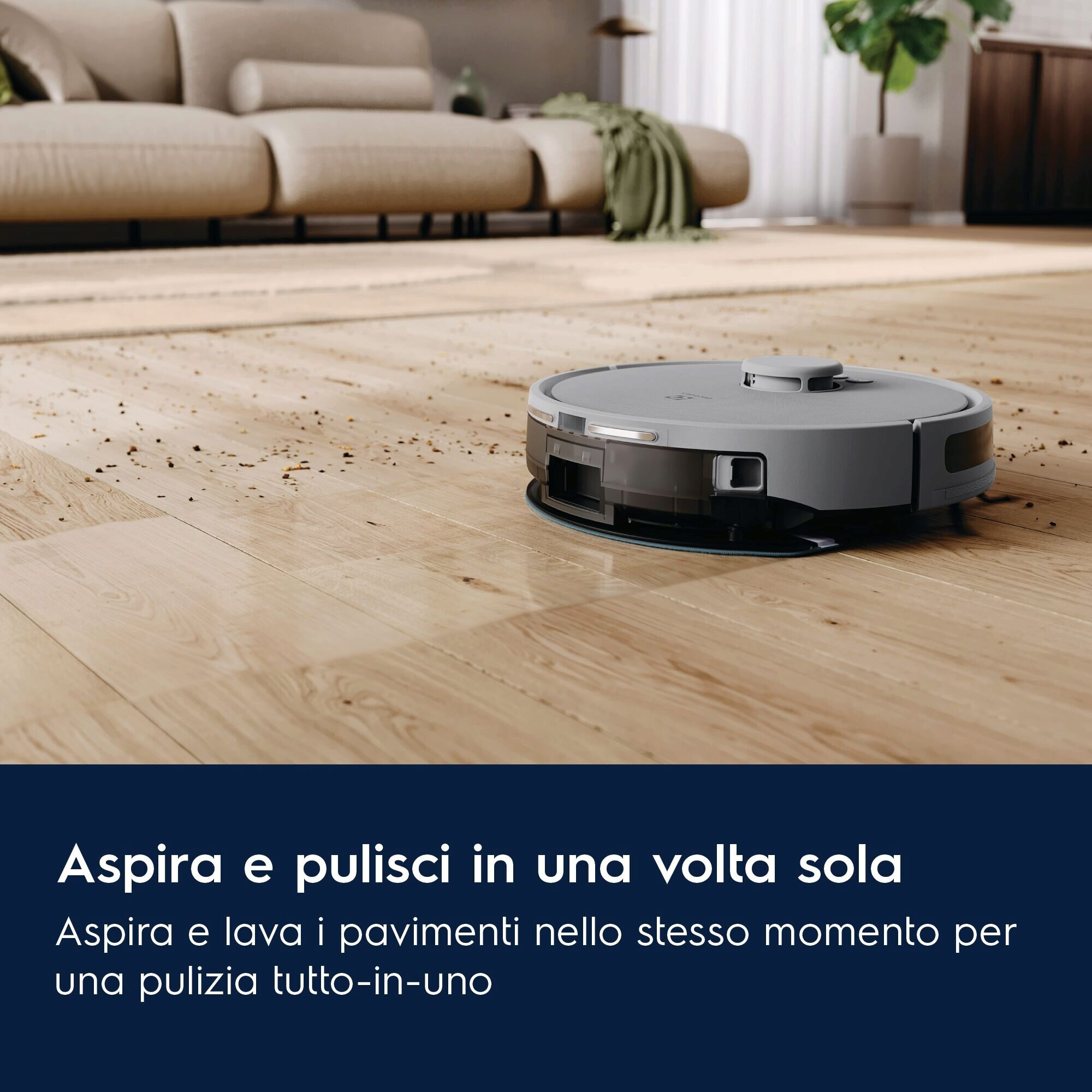 Un aspirapolvere robot grigio su un pavimento in legno. Un divano e una pianta sullo sfondo. Il pavimento è sporco.