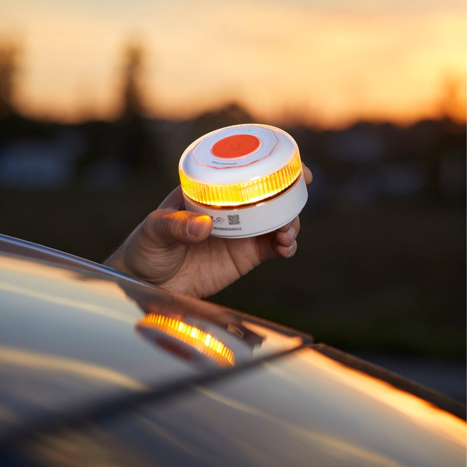 Una mano sostiene una luz de emergencia blanca y naranja que emite luz naranja. El fondo muestra una puesta de sol.