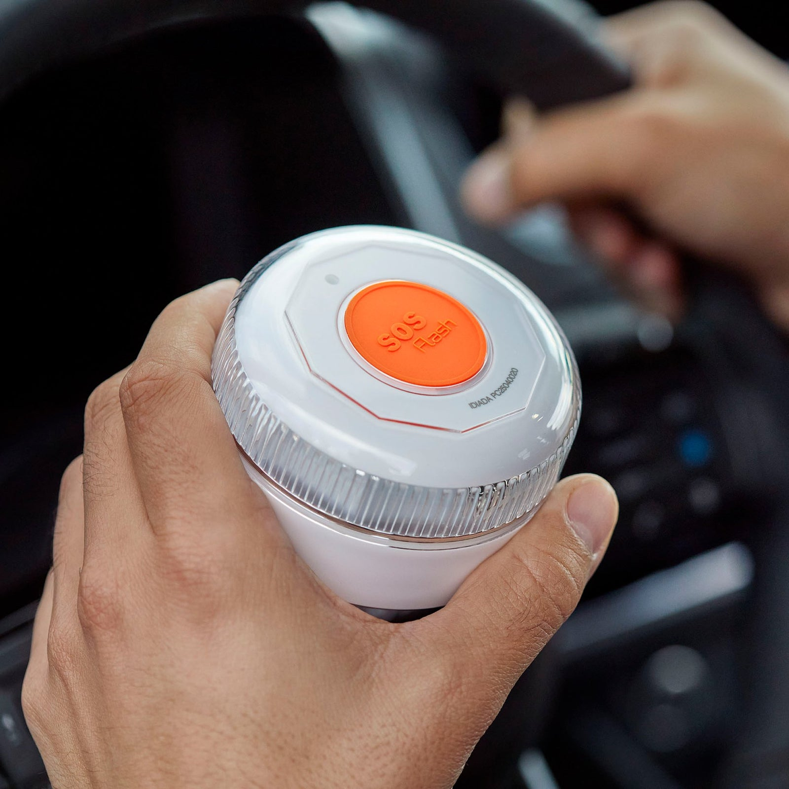 Una persona sostiene un dispositivo de flash SOS blanco y naranja en un coche. El dispositivo es redondo, el fondo está borroso.