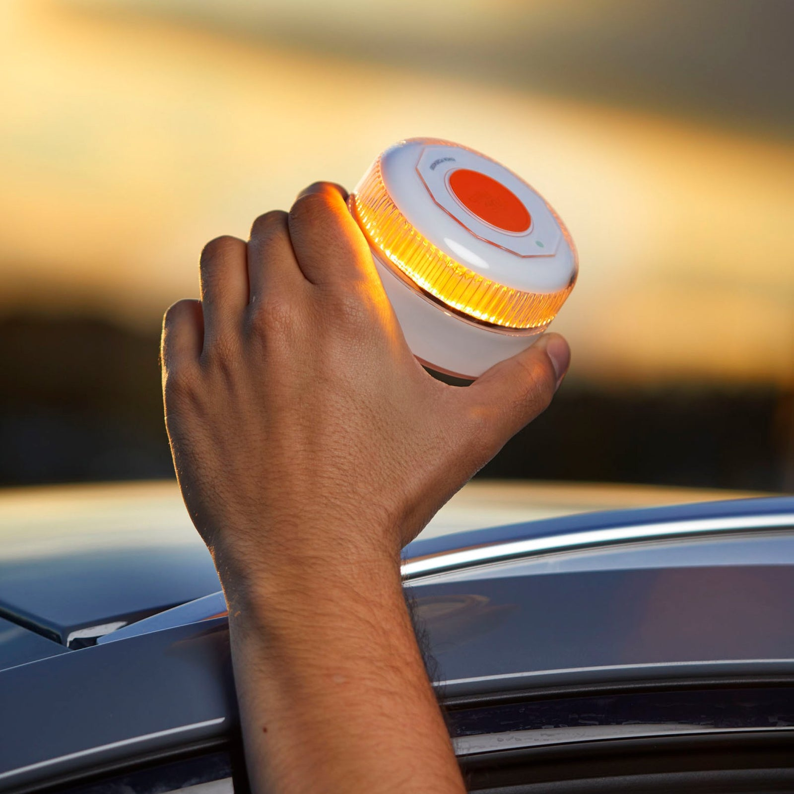 Una mano sostiene una luz de emergencia blanca y naranja. La luz está brillando. El coche es gris. El fondo está borroso.
