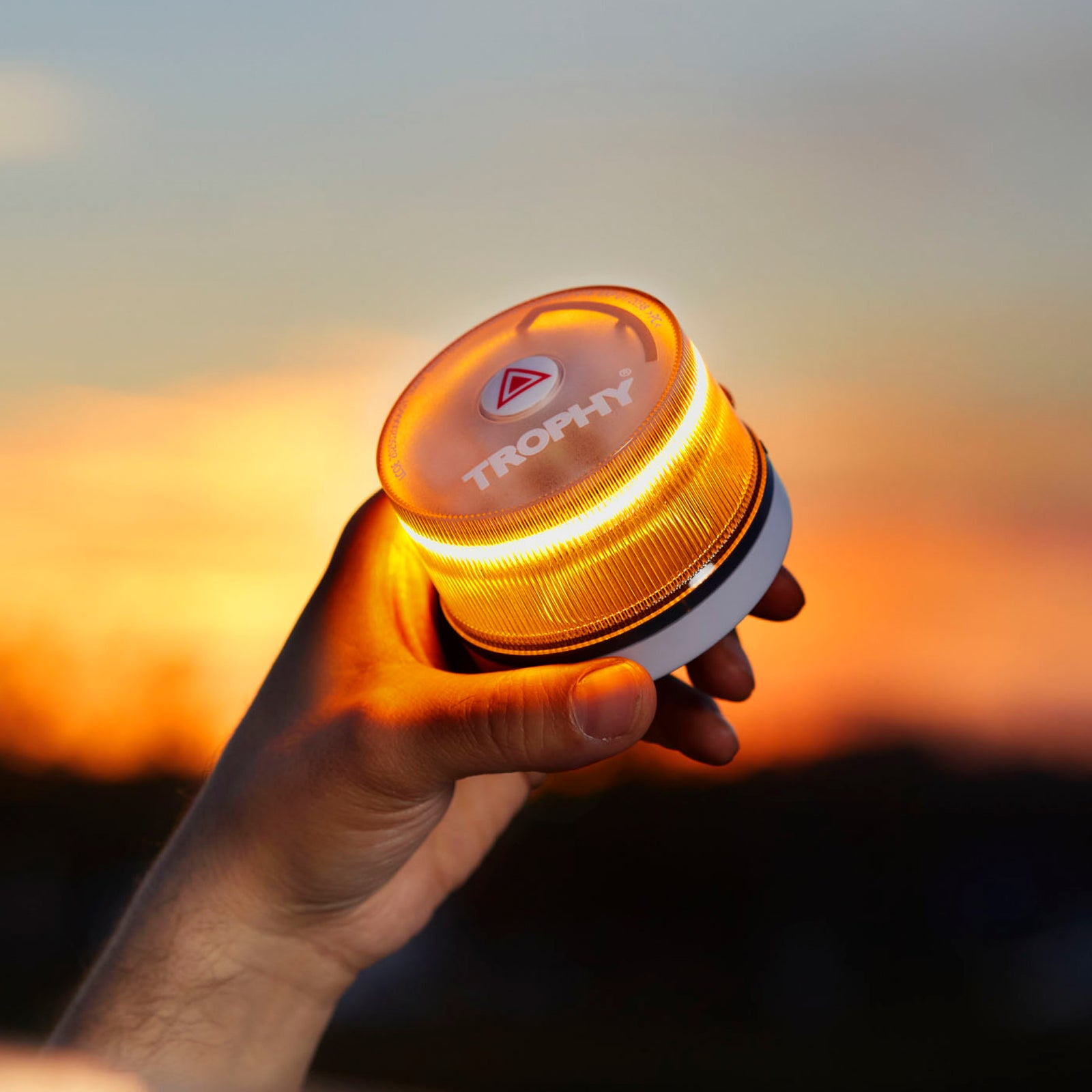 Mano sosteniendo una luz de seguridad redonda de color naranja-amarillo encendida contra un cielo al atardecer. La luz está etiquetada como 'TROPHY'.