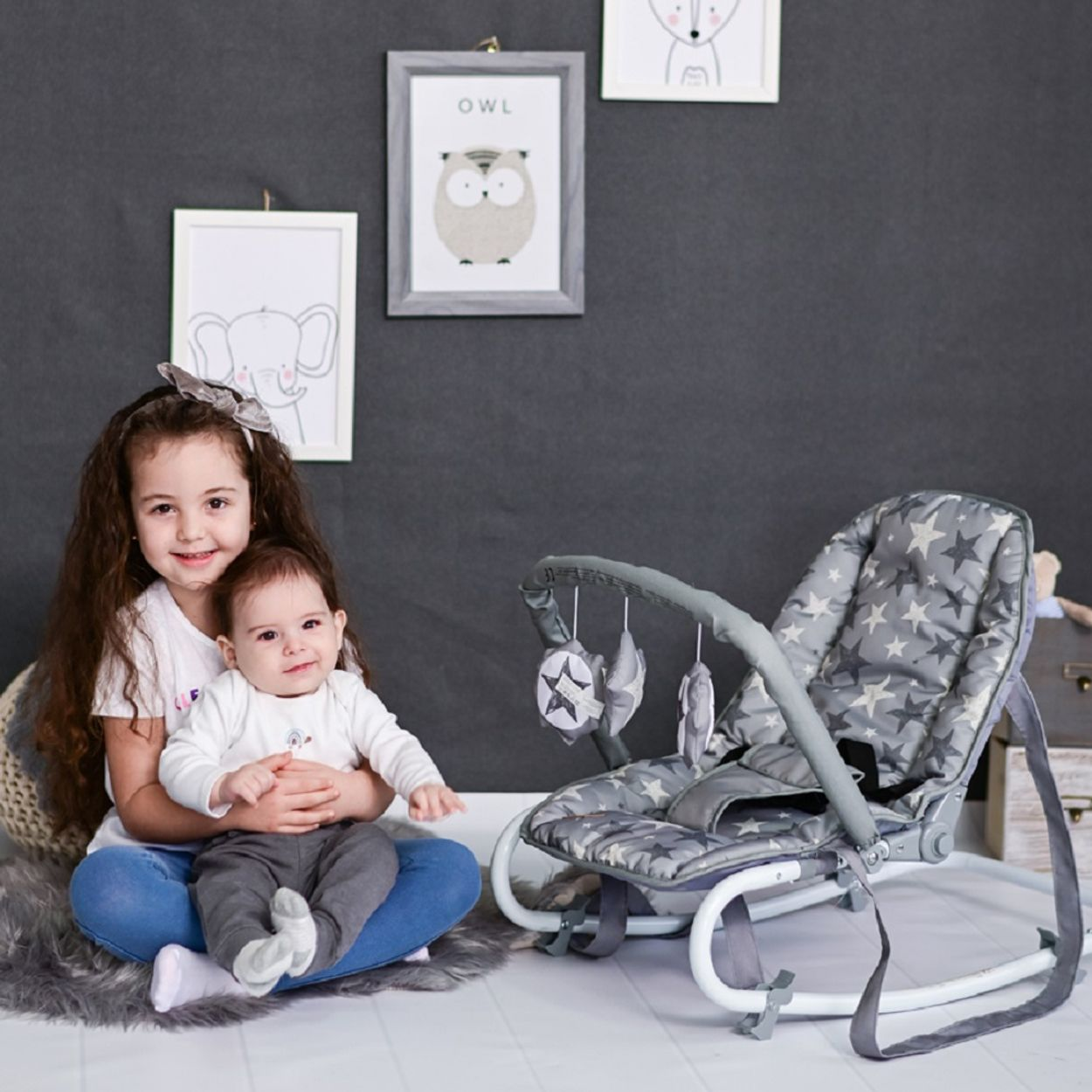 Zwei Kinder sitzen auf dem Boden. Eines hält ein Baby. Neben ihnen steht eine graue Babywippe mit Sternenmuster. Bilder hängen an der Wand.