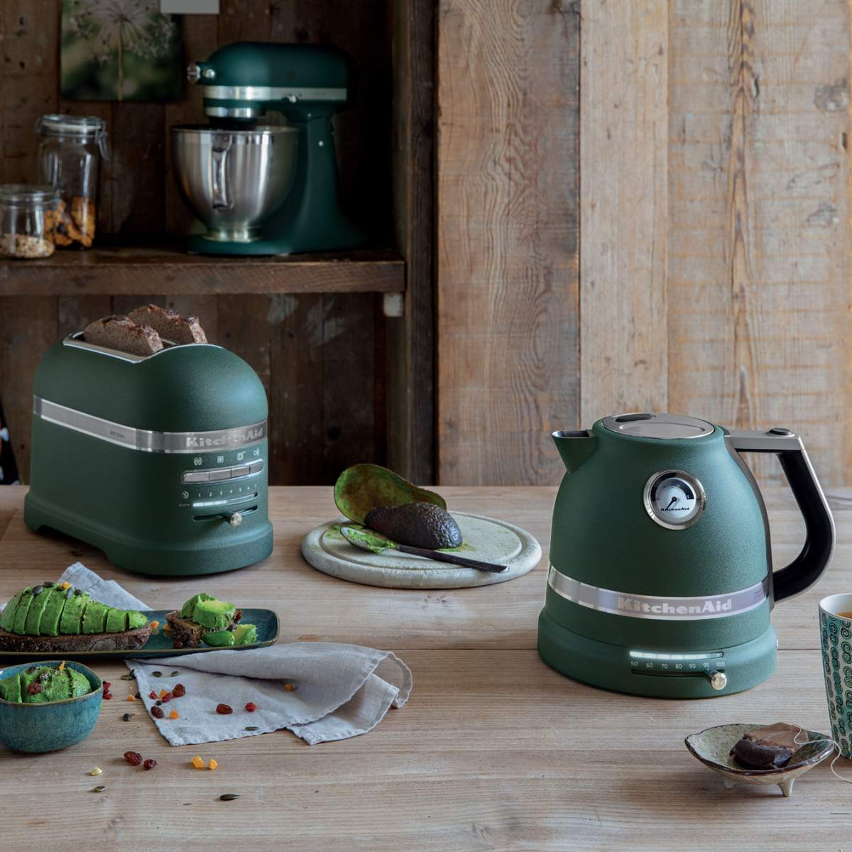 Eine Küchenszene mit grünen Geräten: Toaster, Wasserkocher und Küchenmaschine. Avocadotoast und halbierte Avocado auf einem Holztisch.
