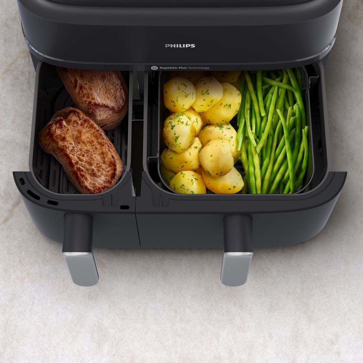 Een zwarte Philips airfryer met steak, aardappelen en sperziebonen. De airfryer heeft twee compartimenten.