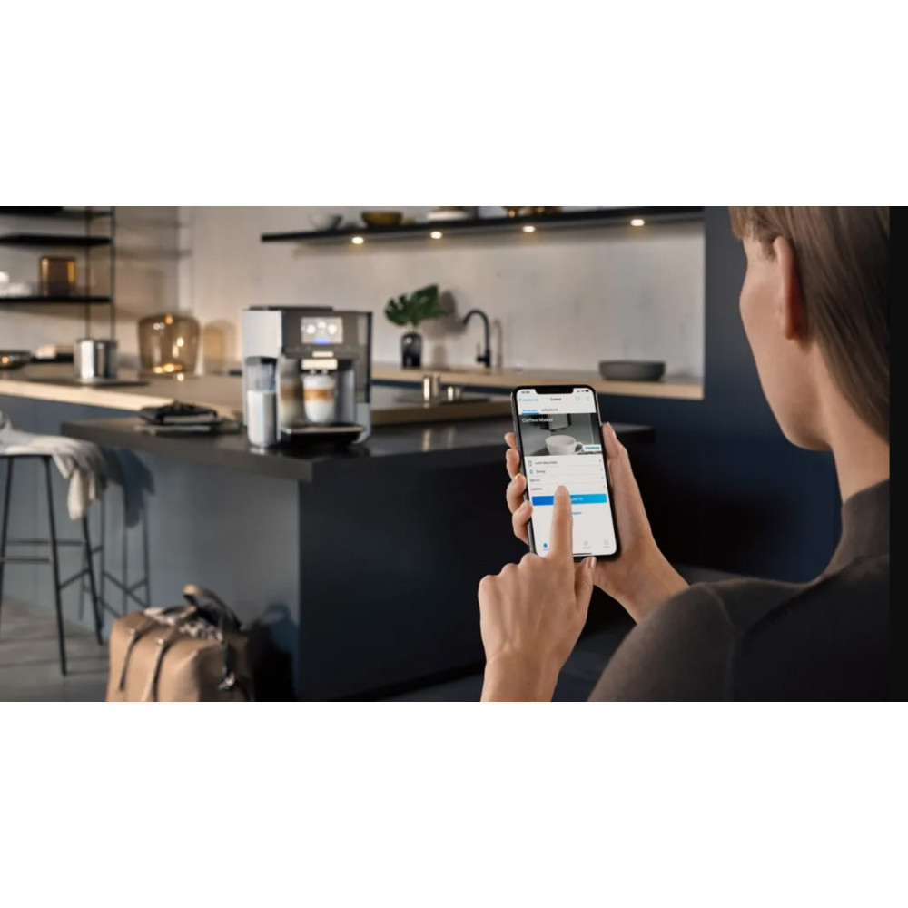 Kobieta używa smartfona do sterowania ekspresem do kawy w nowoczesnej kuchni. Filiżanka kawy jest gotowa. Walizka leży na podłodze.