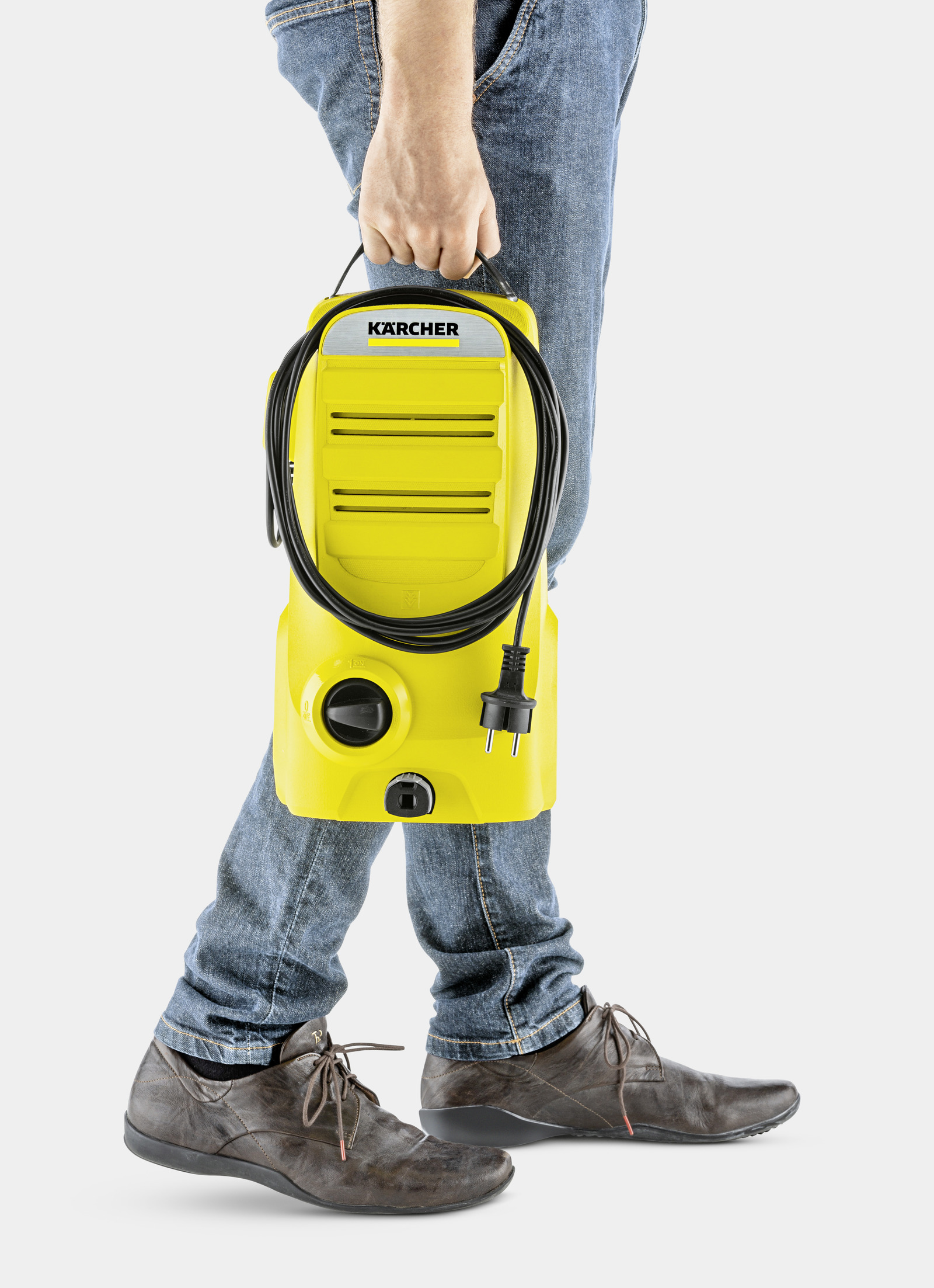 Eine Person in Jeans und braunen Schuhen trägt einen gelben Karcher-Hochdruckreiniger. Schwarzes Kabel und Stecker sind sichtbar.