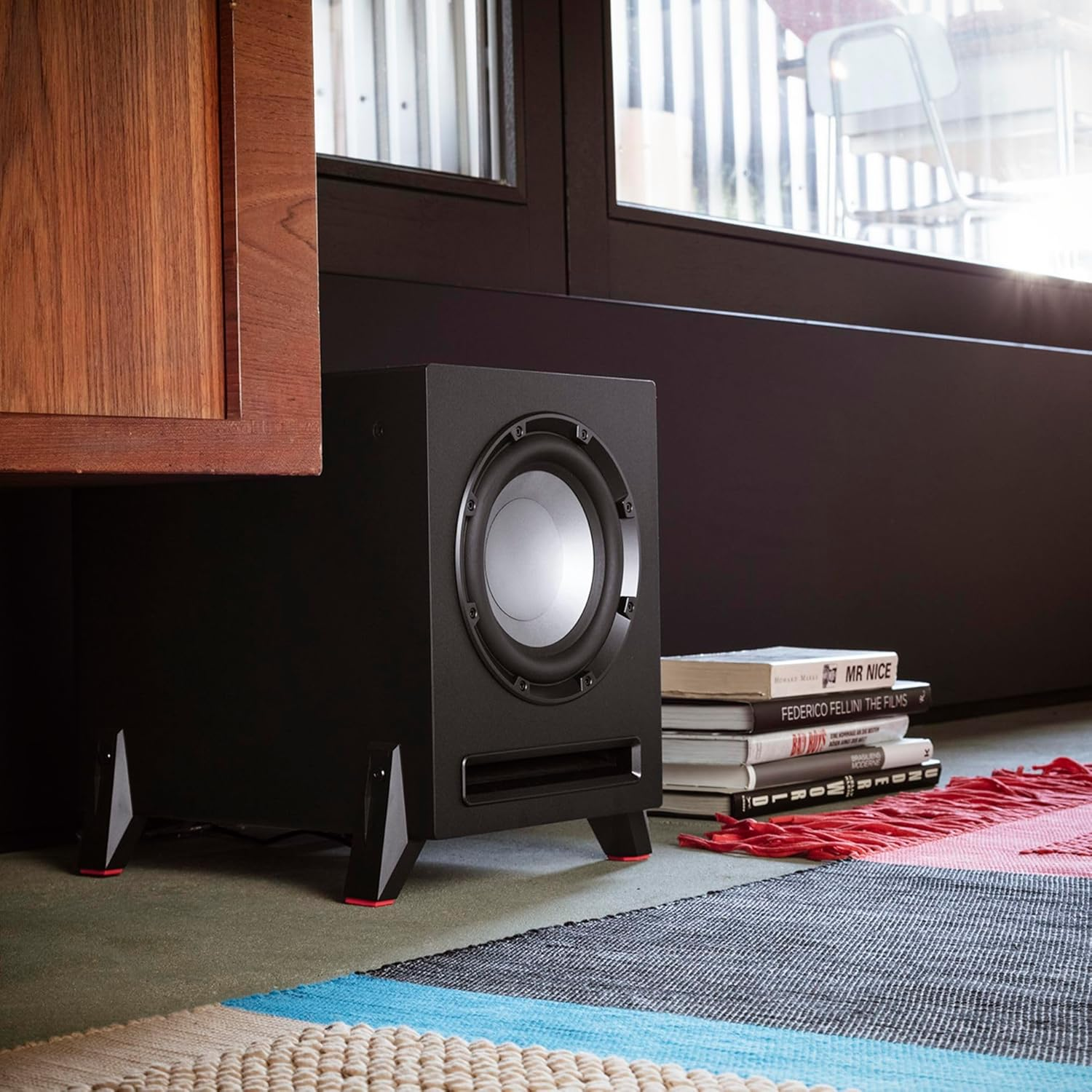 Ein schwarzer Subwoofer steht in der Nähe eines Fensters. Bücher und ein bunter Teppich liegen auf dem Boden. Links ein Holzschrank.